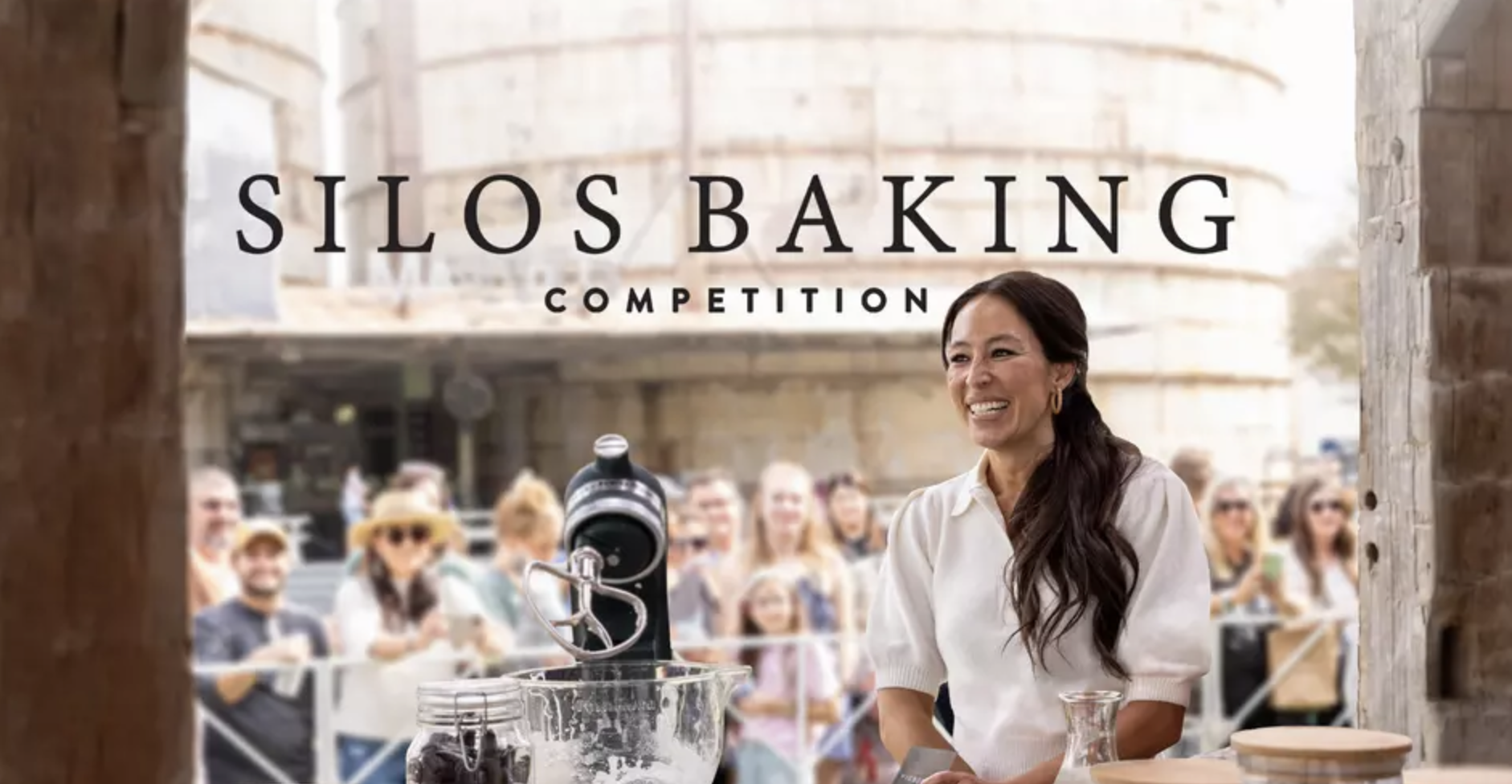 A woman at a silos baking competition, smiling and standing in front of a crowd outdoors. There is a large sign reading 'Silos Baking Competition' behind her.