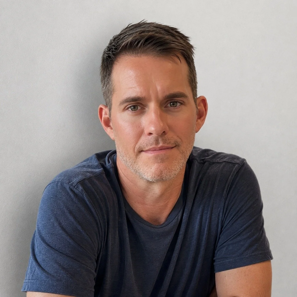 A man with short dark hair, stubble, and a slight smile, wearing a navy T-shirt, sitting against a light gray wall.