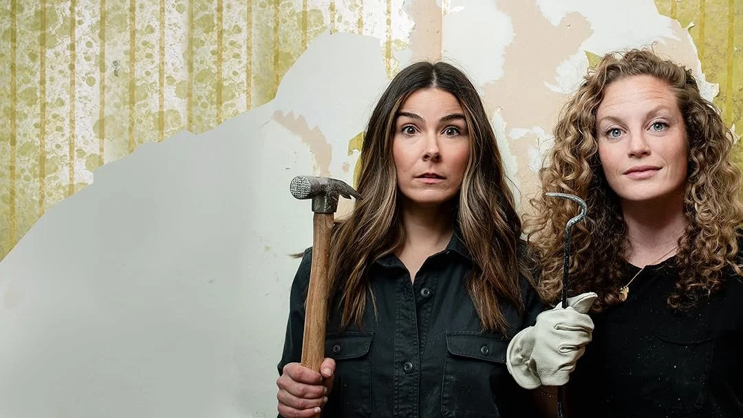 Two women with curious expressions standing in front of a peeling wallpaper wall, one holding a hammer and the other holding a curved tool.