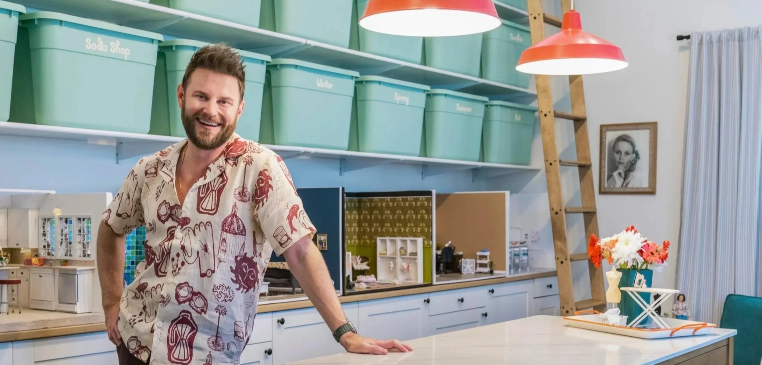A man with a beard smiling, standing in a colorful room with shelves labeled Soda Shop, Water, Spring, Summer, and Fall, with a table in front and a framed black and white portrait on the wall.