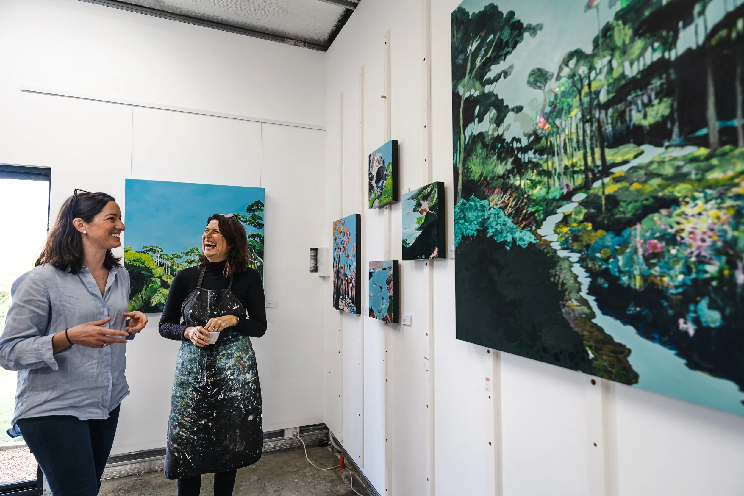 Two women are standing and talking in an art gallery. They are smiling and seem to be enjoying their conversation. The gallery walls are decorated with colorful landscape paintings.