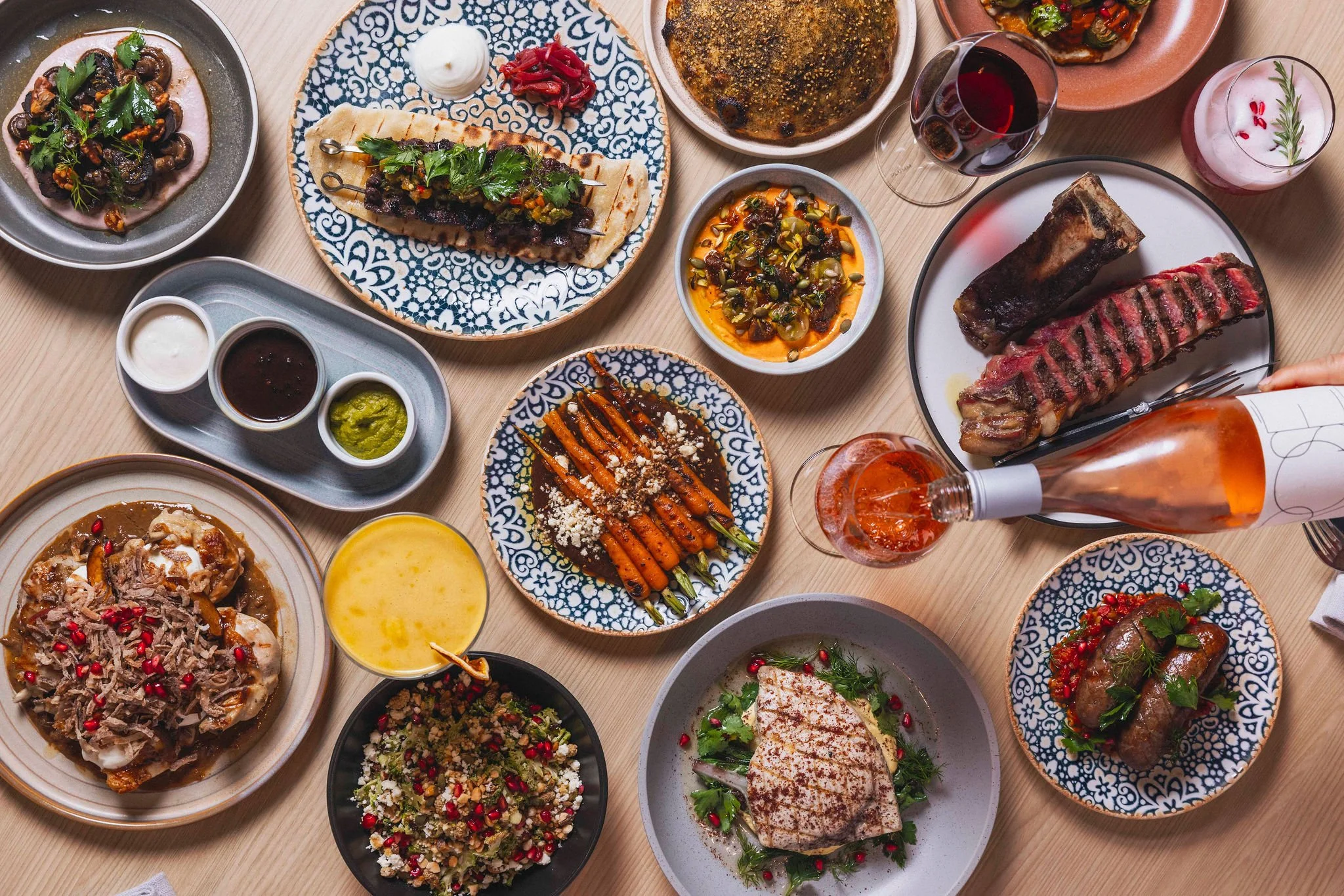 A table filled with a variety of Mexican dishes, including grilled meats, tacos, salads, and sauces, with wine and beverages.