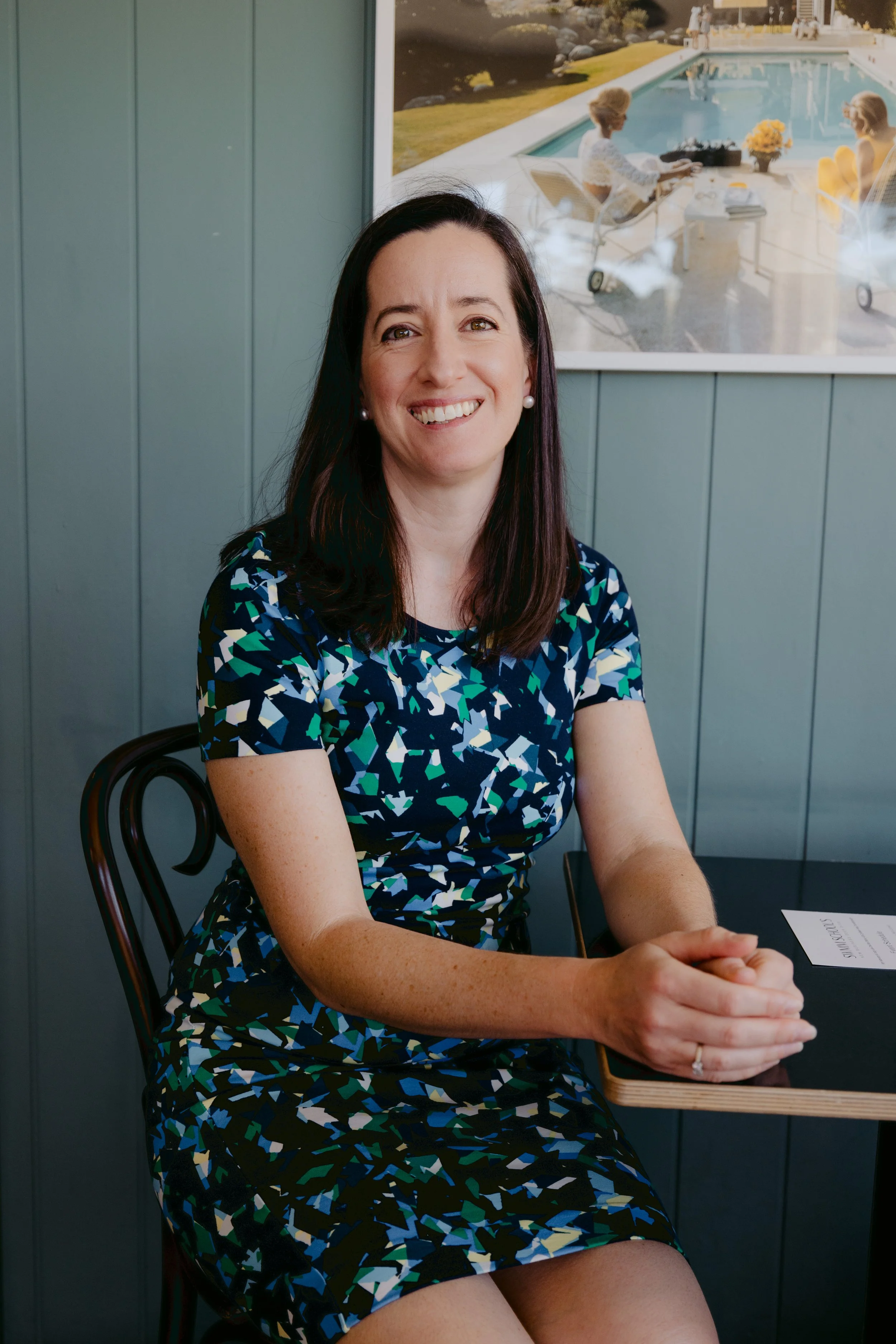 A woman with long dark hair, wearing pearl earrings and a patterned dress in blue, black, white, and green, sitting at a table with her hands clasped. She is smiling and looking at the camera, with a light blue wall and a photo of people by a pool in the background.