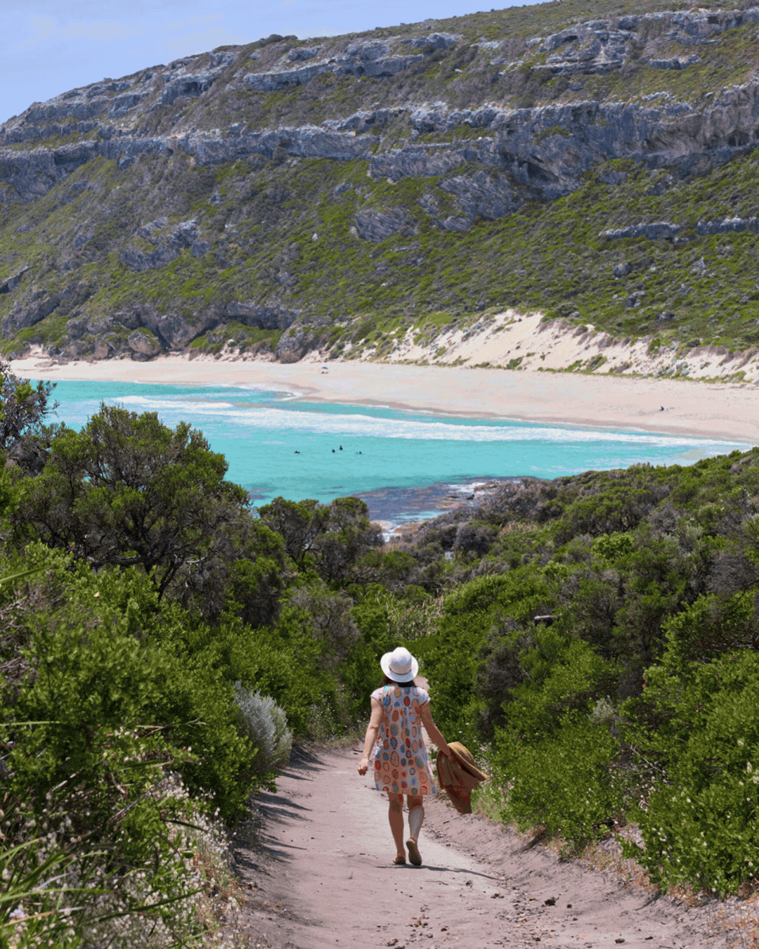 A person wearing a sunhat and a patterned dress walks down a sandy trail through green shrubbery toward a beach with turquoise water and large rocky cliffs in the background.