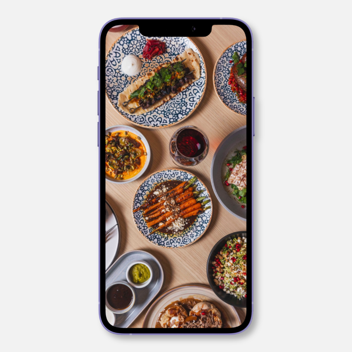 Smartphone displaying a top-down photo of a table with various Middle Eastern dishes on patterned plates, including vegetables, hummus, a salad, and a glass of red wine.