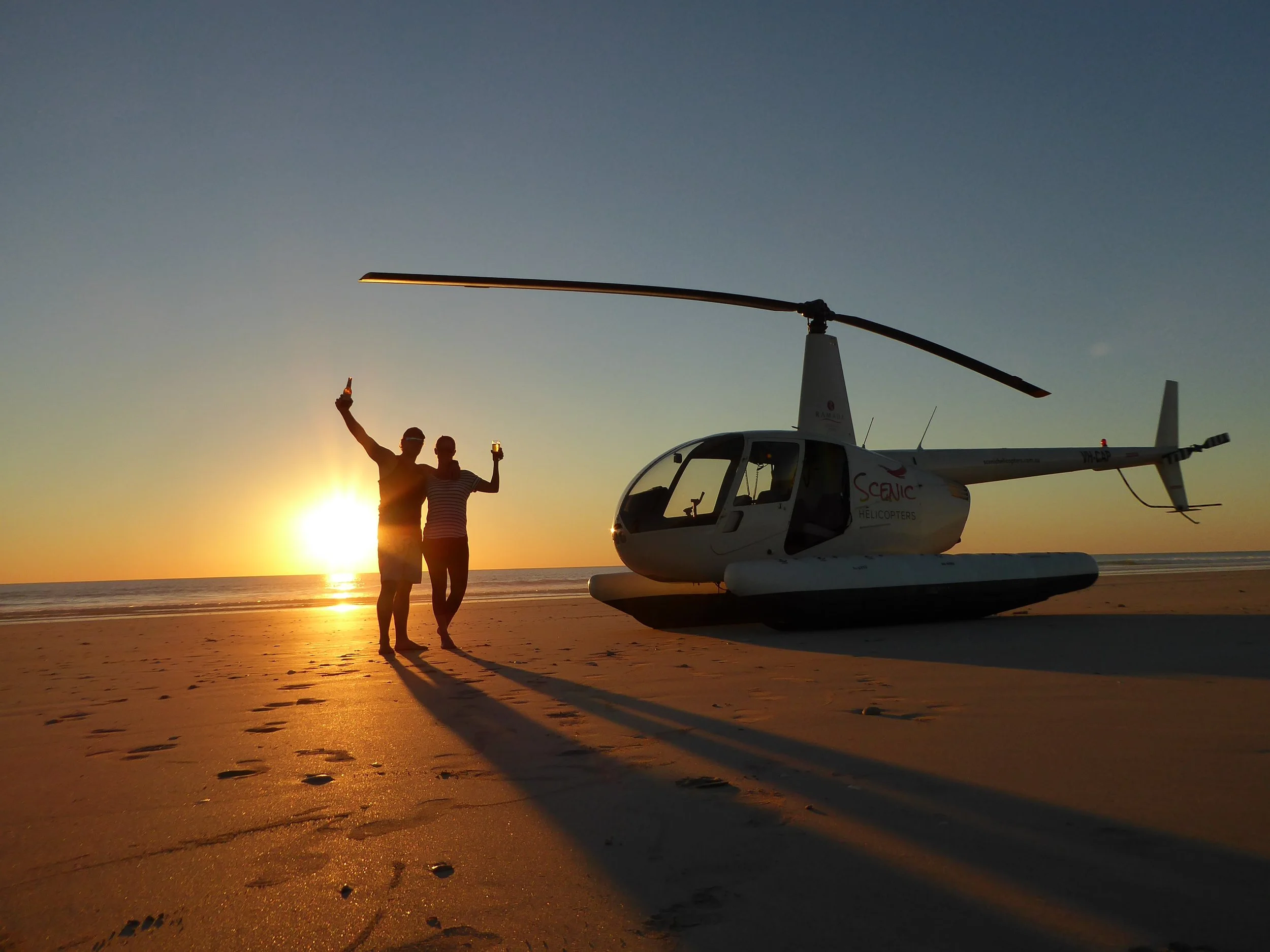 Two people on a beach at sunset taking a selfie beside a white helicopter with a rotor blade on top, the sun setting behind them, casting long shadows on the sand.