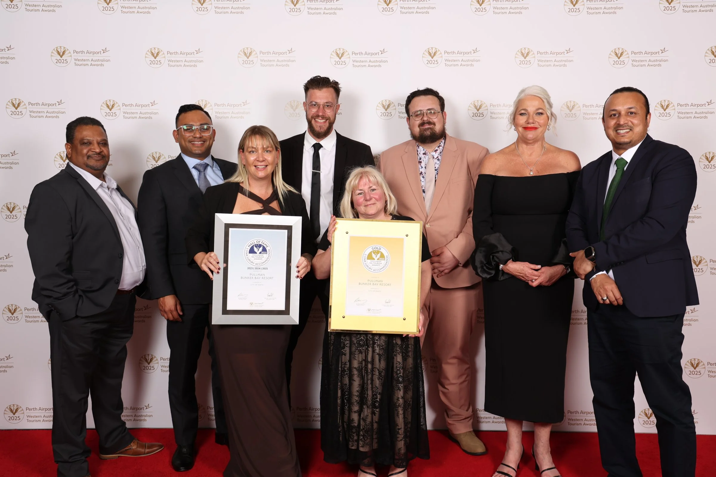 A group of nine people dressed in formal attire standing in front of a white backdrop with the logos of Perth Airport and the Western Australian Tourism Awards, holding certificates and awards.