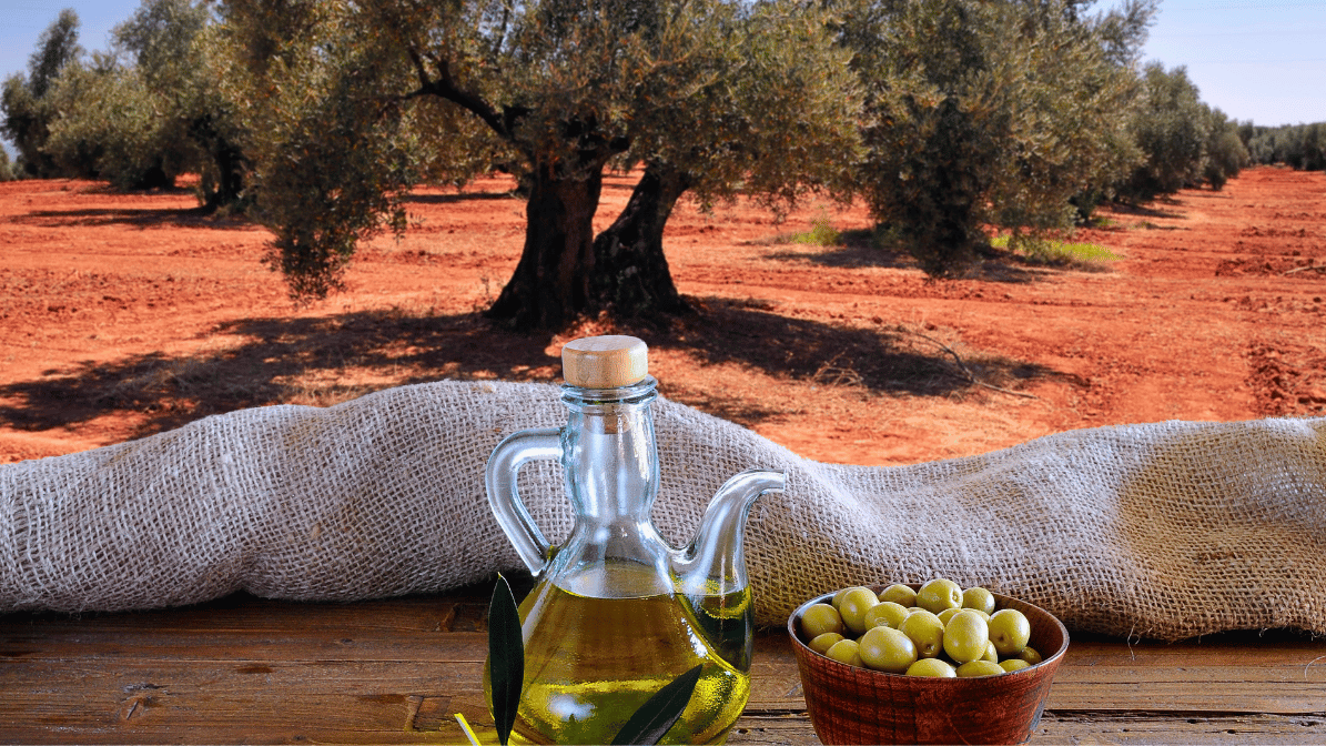 OliveByNature Olive oil in a glass bottle with a cork, green olives in a bowl on a wooden surface, and a burlap cloth with a backdrop of an olive grove with trees and red soil.