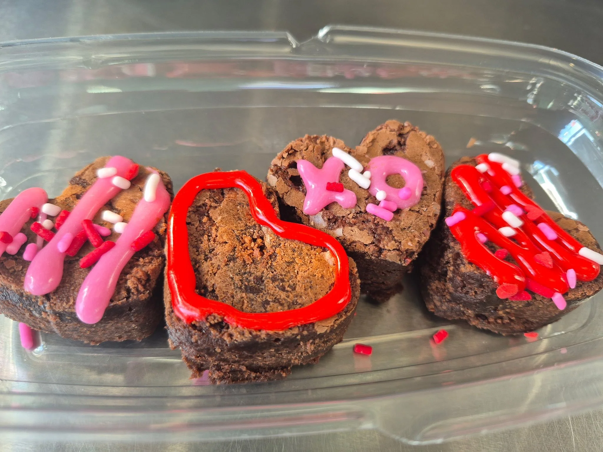 Four heart-shaped chocolate brownies decorated with pink and red icing, sprinkles, and a red heart outline, in a clear plastic container.