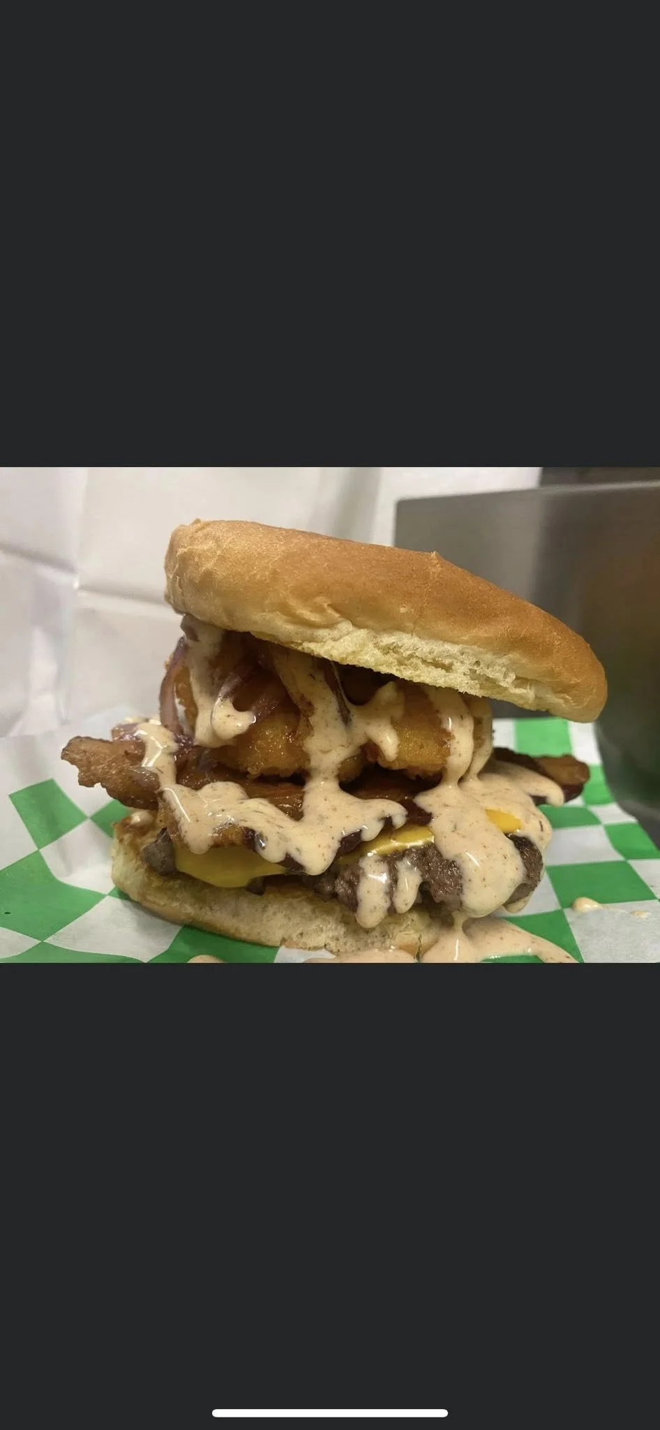A burger with fried chicken, bacon, cheese, and sauce in a bun on checkered paper