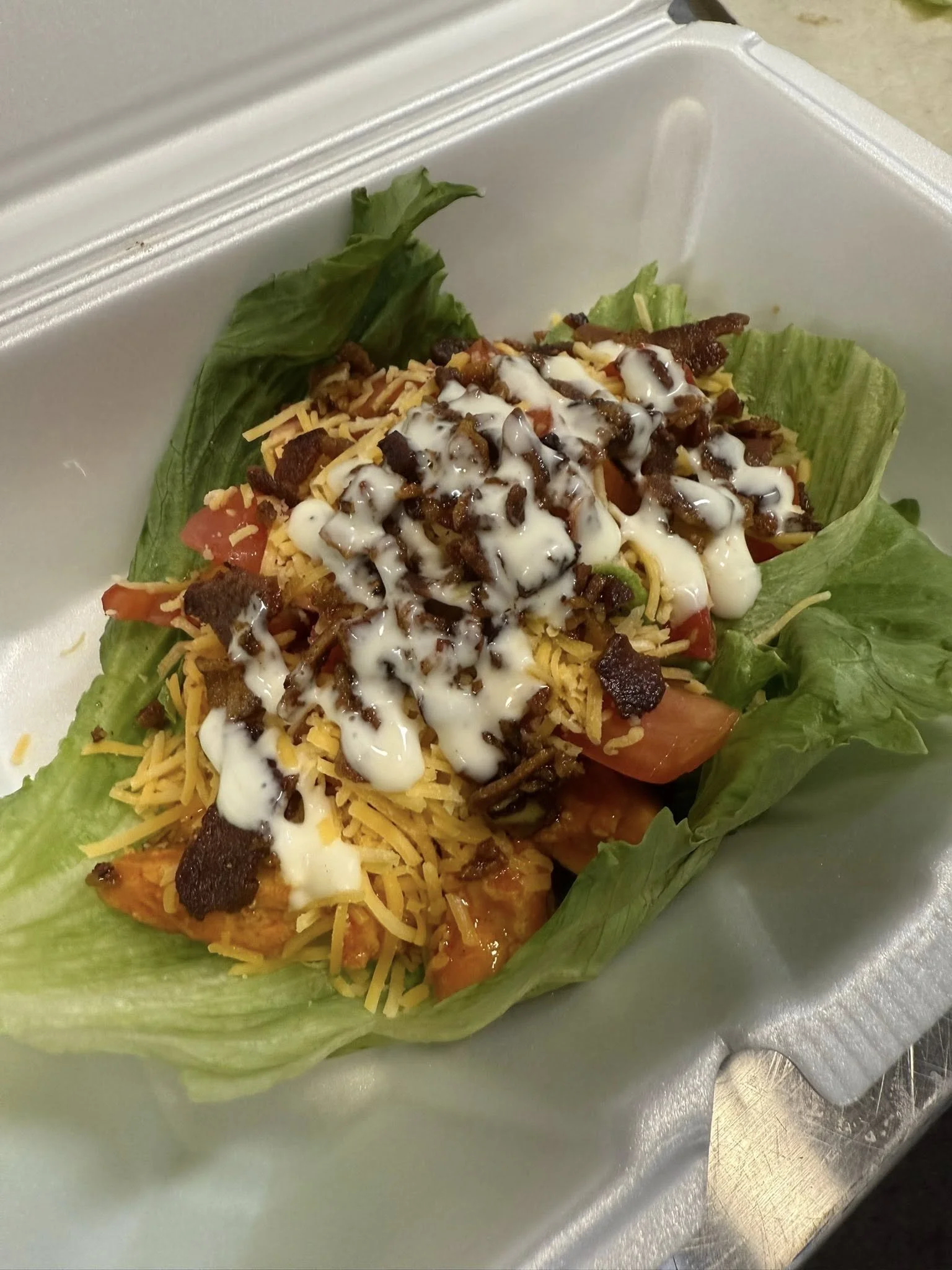 Lettuce leaf with shredded cheese, diced tomatoes, shredded meat, and drizzled white sauce in a foam takeaway container.