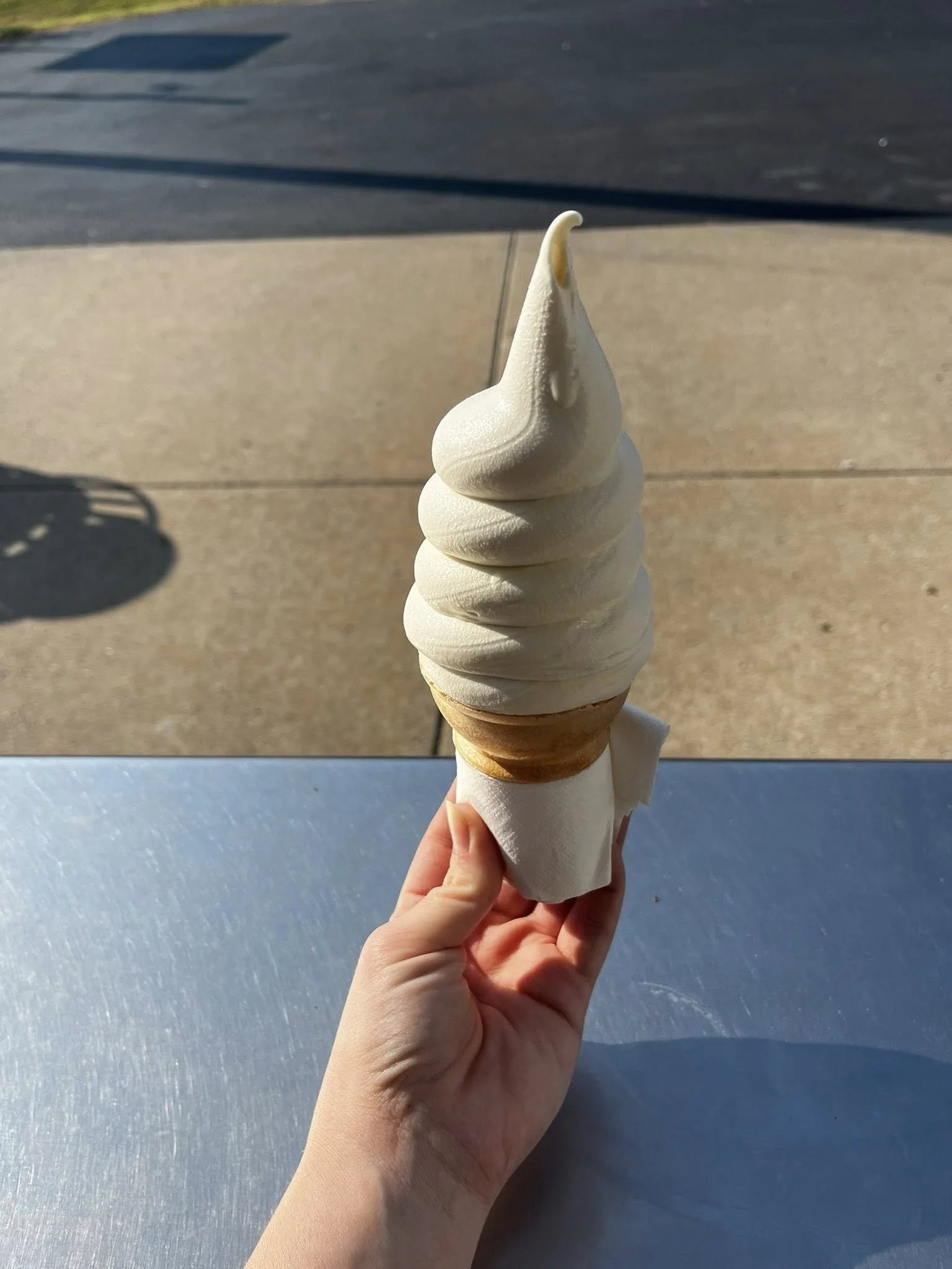 Close-up of a vanilla soft serve ice cream cone held by hand against outdoor setting with concrete and shadow.