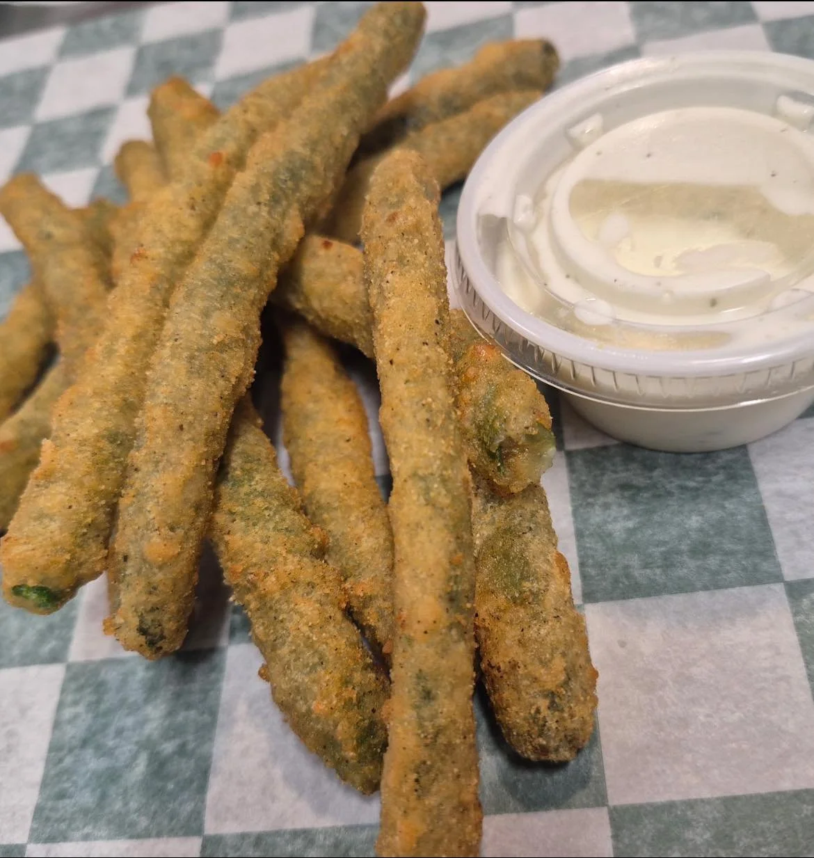Battered fried green beans with a cup of creamy dipping sauce on checkered paper.