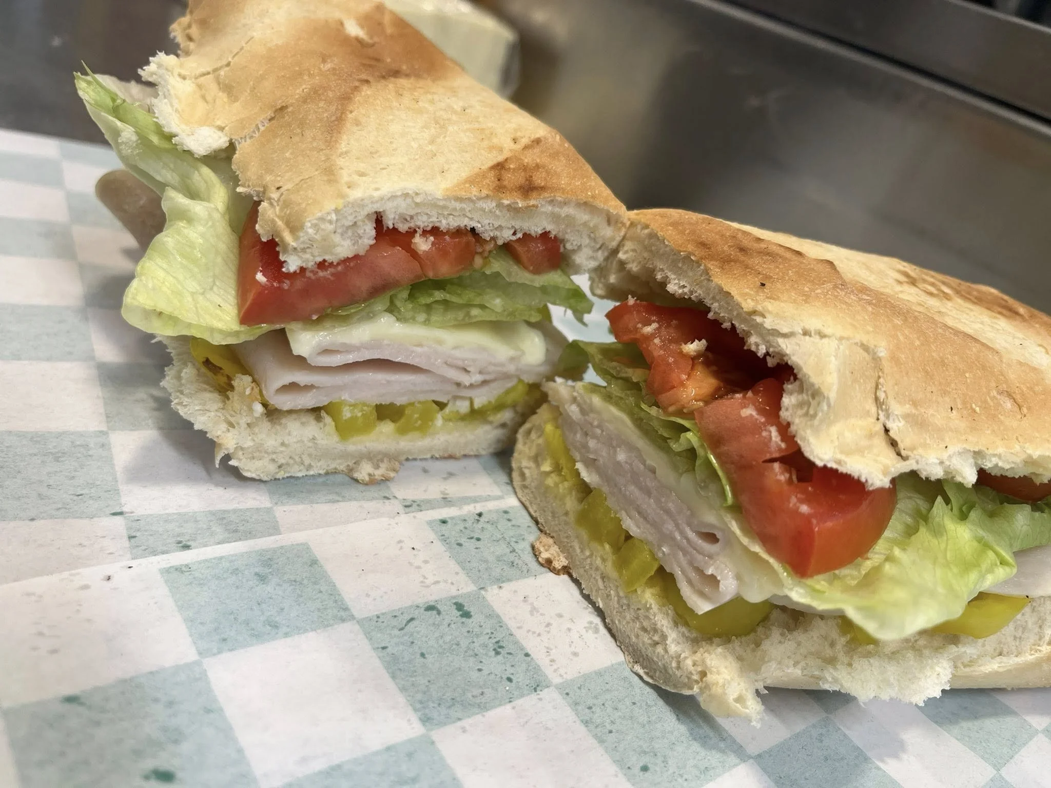 A sandwich with lettuce, tomato, pickles, turkey slices, and mayonnaise in a toasted hoagie bun on checkered paper.