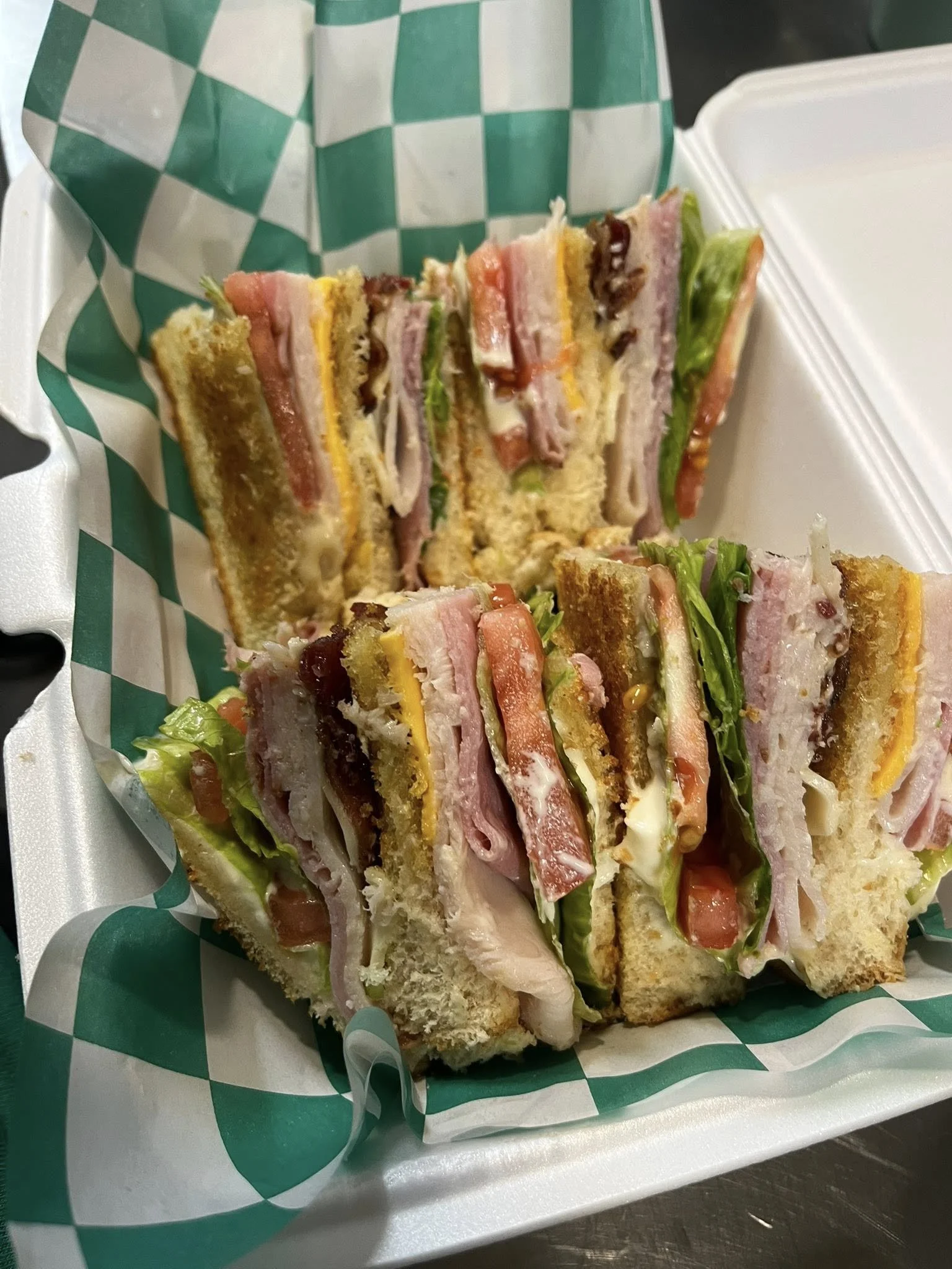 A club sandwich with layers of turkey, bacon, lettuce, tomato, cheese, and mayonnaise, cut into quarters and served in a takeaway container lined with green and white checkered paper.