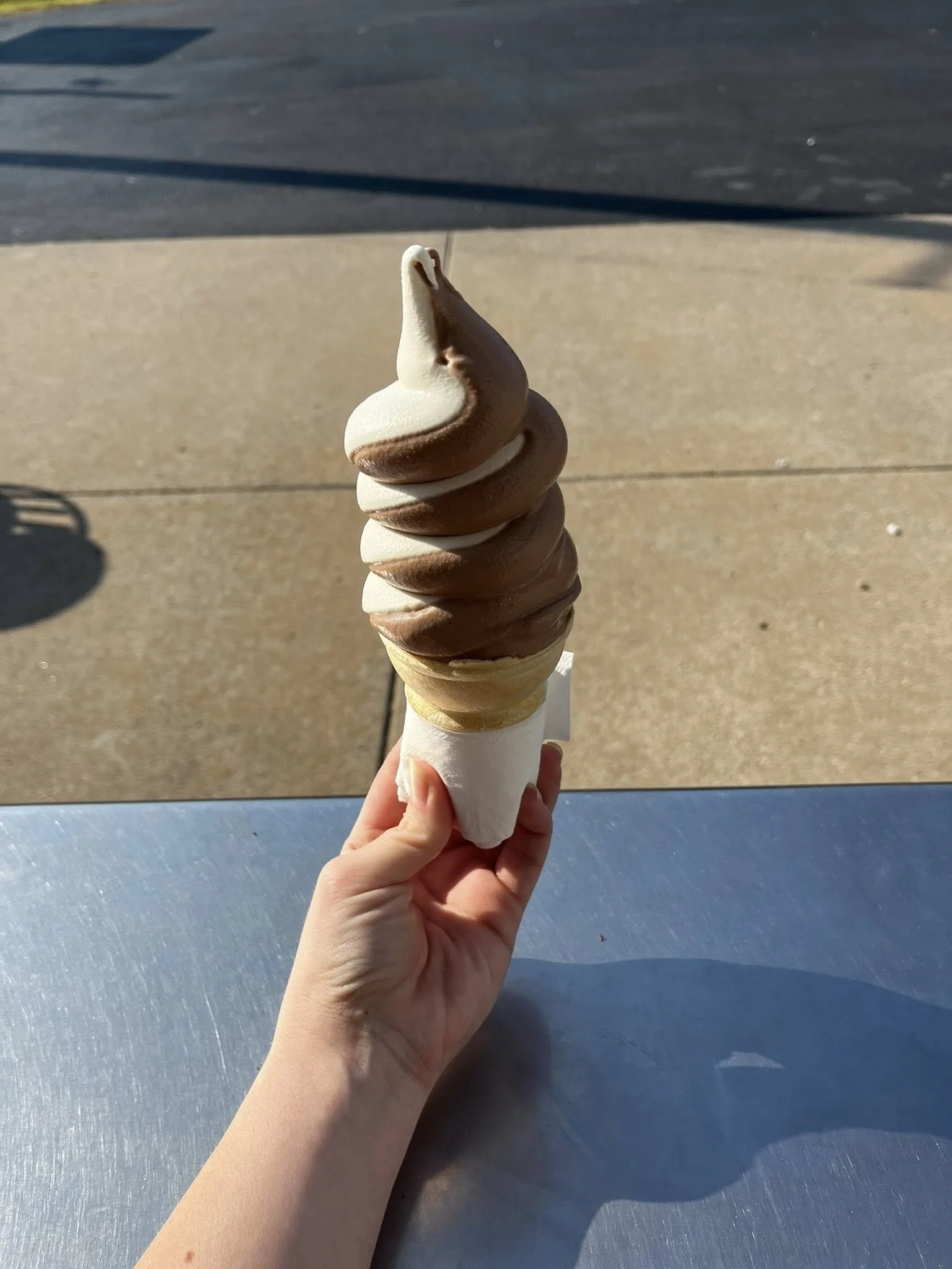 A hand holding a large soft serve ice cream cone with swirls of chocolate and vanilla ice cream, outdoors on a sunny day.