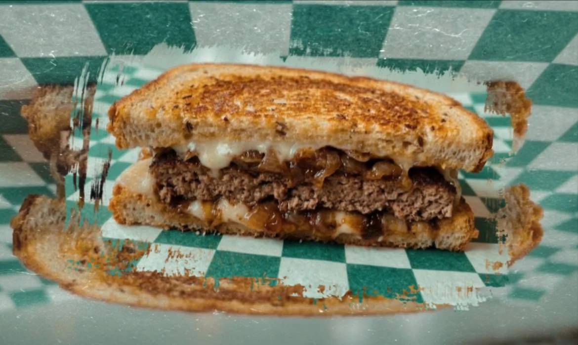 Close-up of a toasted sandwich with melted cheese, cooked ground beef, and caramelized onions inside, on green checkered paper.