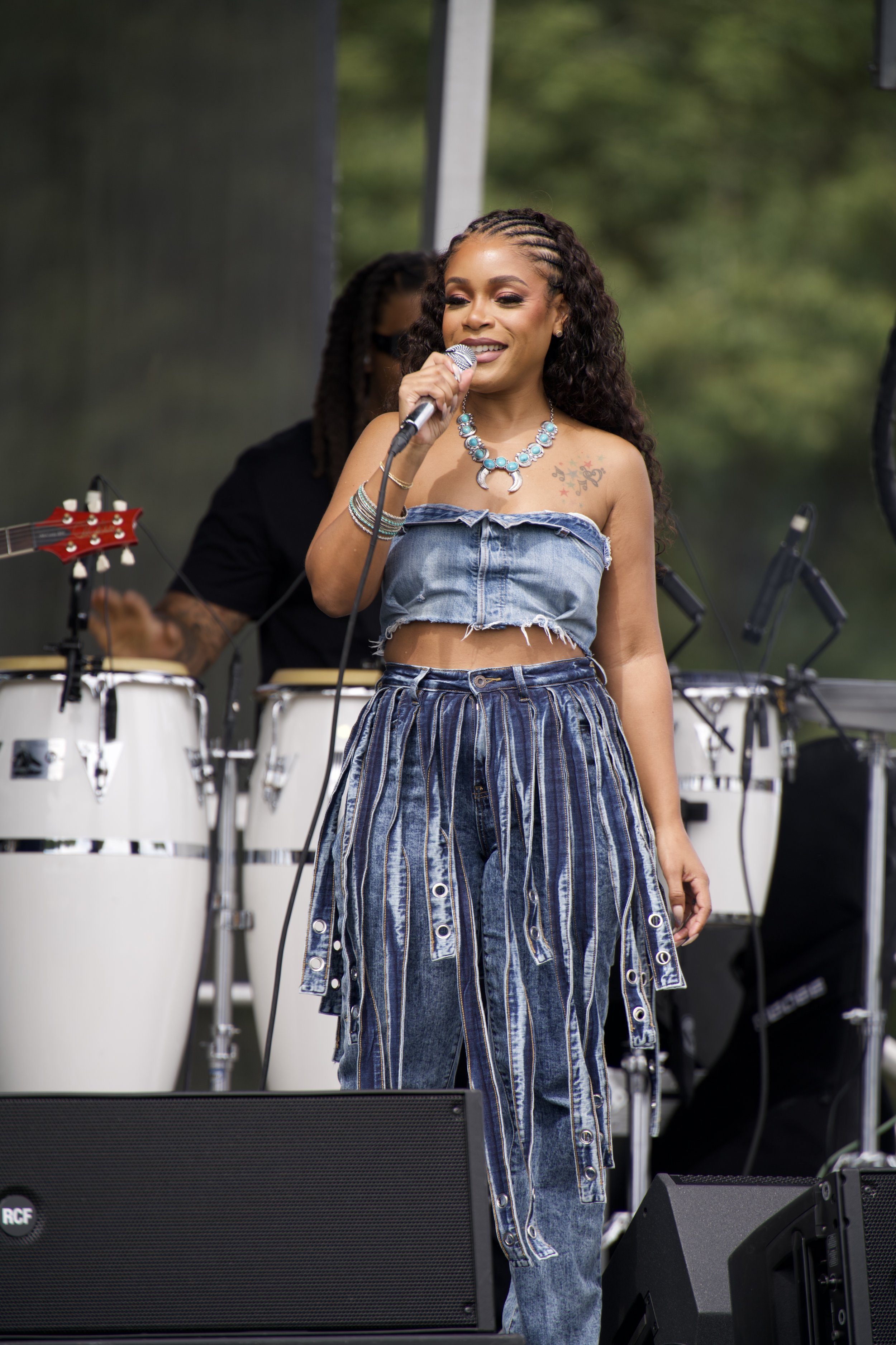 A woman singing into a microphone on stage during a live outdoor performance, with a musician playing percussion instruments behind her.