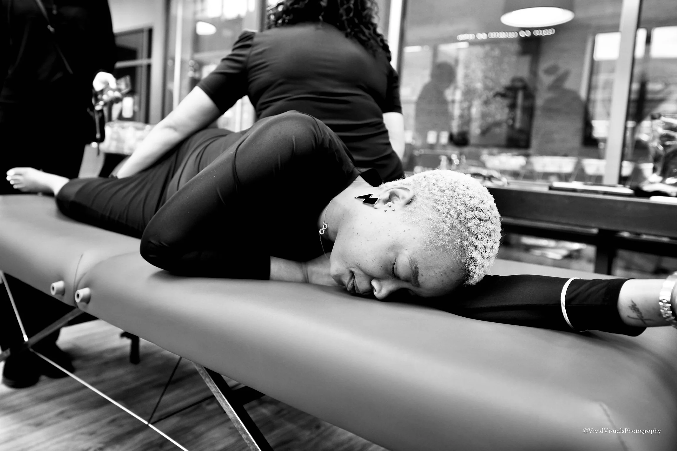 A woman with short, curly hair lying face down on a massage table, receiving a massage. She has earrings and a necklace. The scene appears to be in a spa or massage clinic with glass windows and glowing lights in the background.