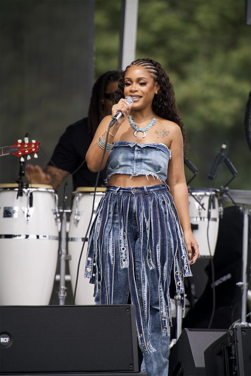 A woman singing into a microphone on stage, wearing a denim off-shoulder crop top and baggy jeans with vertical stripes. Behind her, musicians play percussion instruments, and trees are visible in the background.