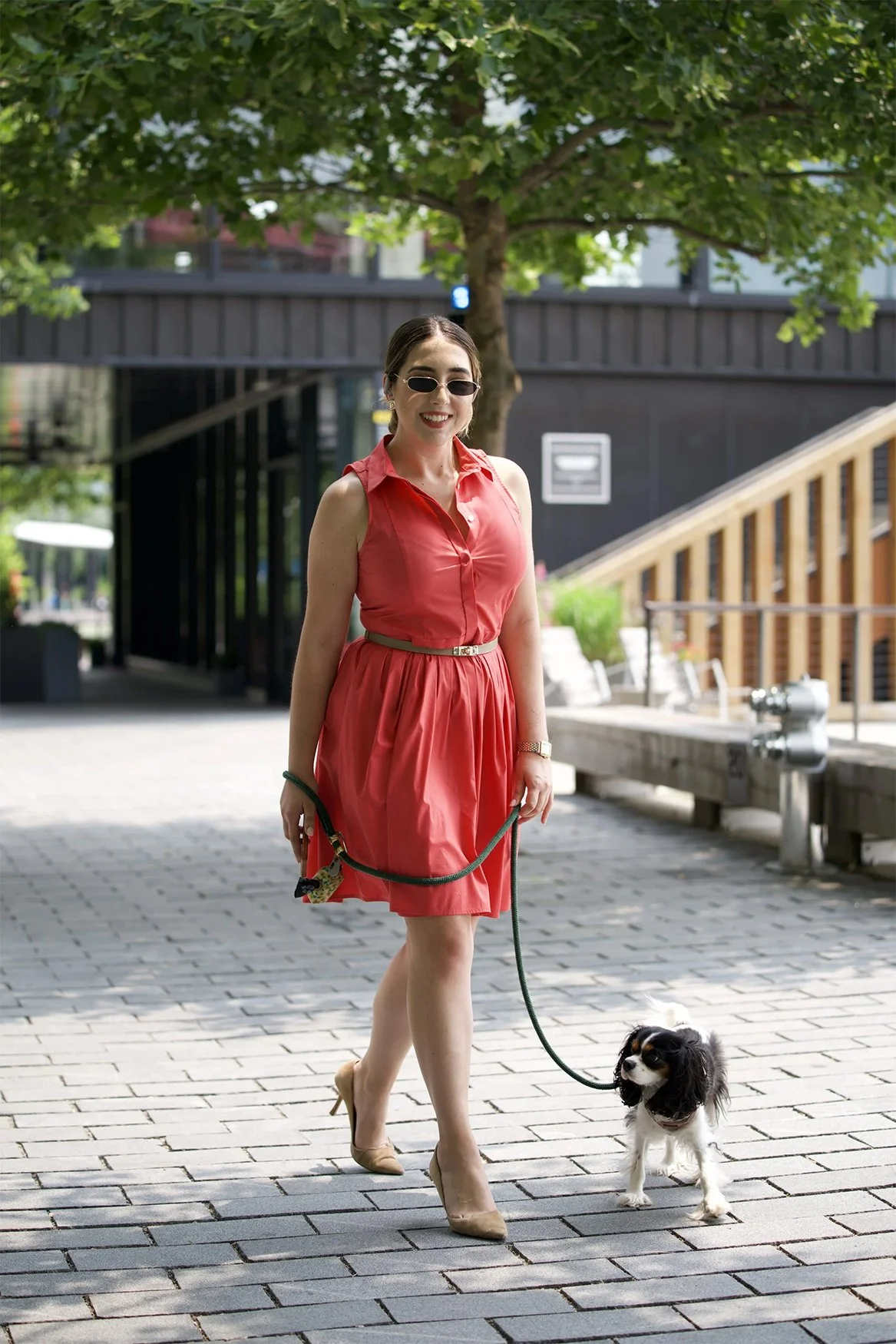 A woman in a coral dress, wearing sunglasses and beige heels, walking a small black and white dog on a leash in an outdoor urban area with trees and modern buildings in the background.