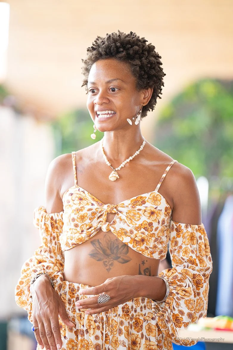 A woman smiling, wearing a floral off-the-shoulder top and matching skirt, with a tattoo visible on her stomach, jewelry, and earrings, standing outdoors with a blurred green background.