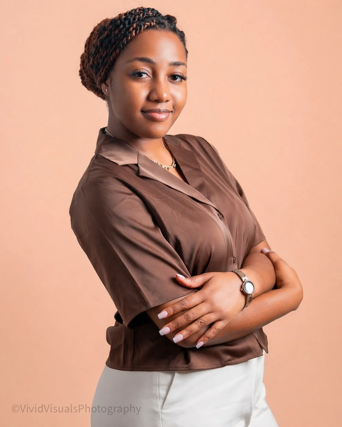 A confident woman with arms crossed, wearing a brown satin shirt, gold necklace, watch, and white pants, standing against a peach background.