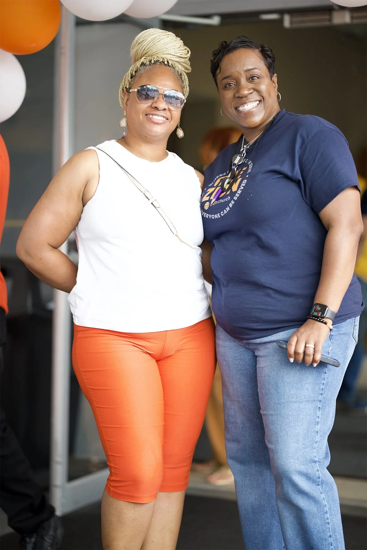 Two women smiling together at an indoor event. One is wearing sunglasses, a white sleeveless top, and orange pants, with her hair styled in a large braid updo. The other is wearing a navy T-shirt, jeans, and a smartwatch, with short black hair.