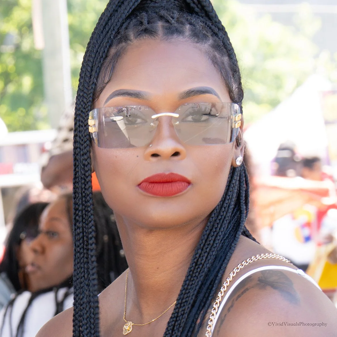 Close-up of a woman with braided hair wearing large sunglasses, red lipstick, a gold necklace, and a white strap top, outdoors with blurred background and people.