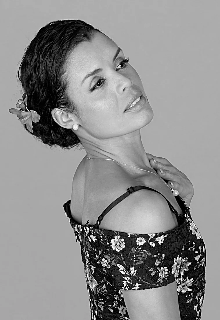 Black and white portrait of a woman with dark, wet-looking hair styled up, wearing floral dress and jewelry, looking slightly upward and to the side with a serene expression.