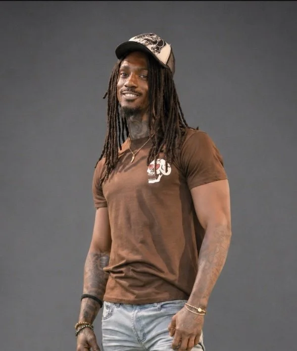 A man with long dreadlocks and tattoos, wearing a brown T-shirt, light jeans, a baseball cap, and jewelry, standing in front of a gray background and smiling.