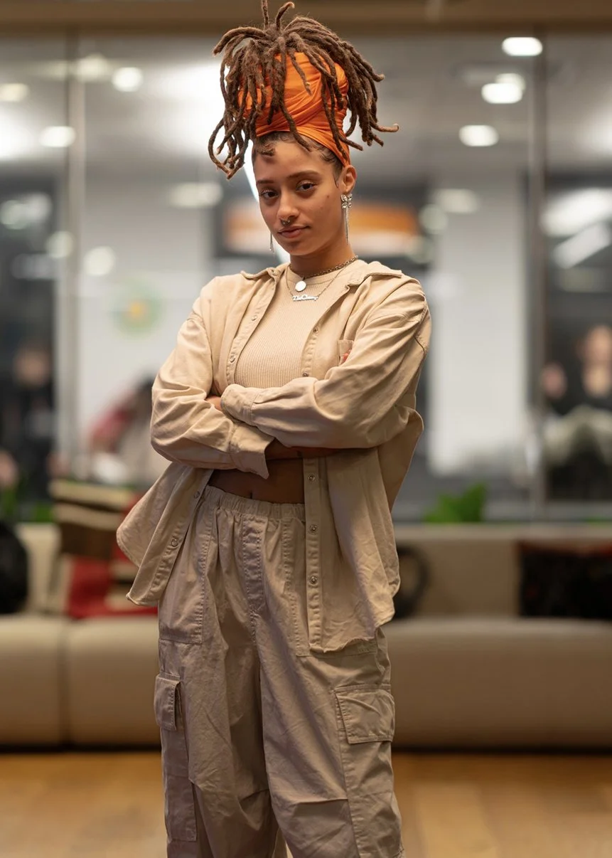 A young woman with dreadlocks wearing an orange head wrap, beige jacket, beige crop top, and cargo pants, standing with arms crossed in an indoor setting, possibly a lounge or waiting area.