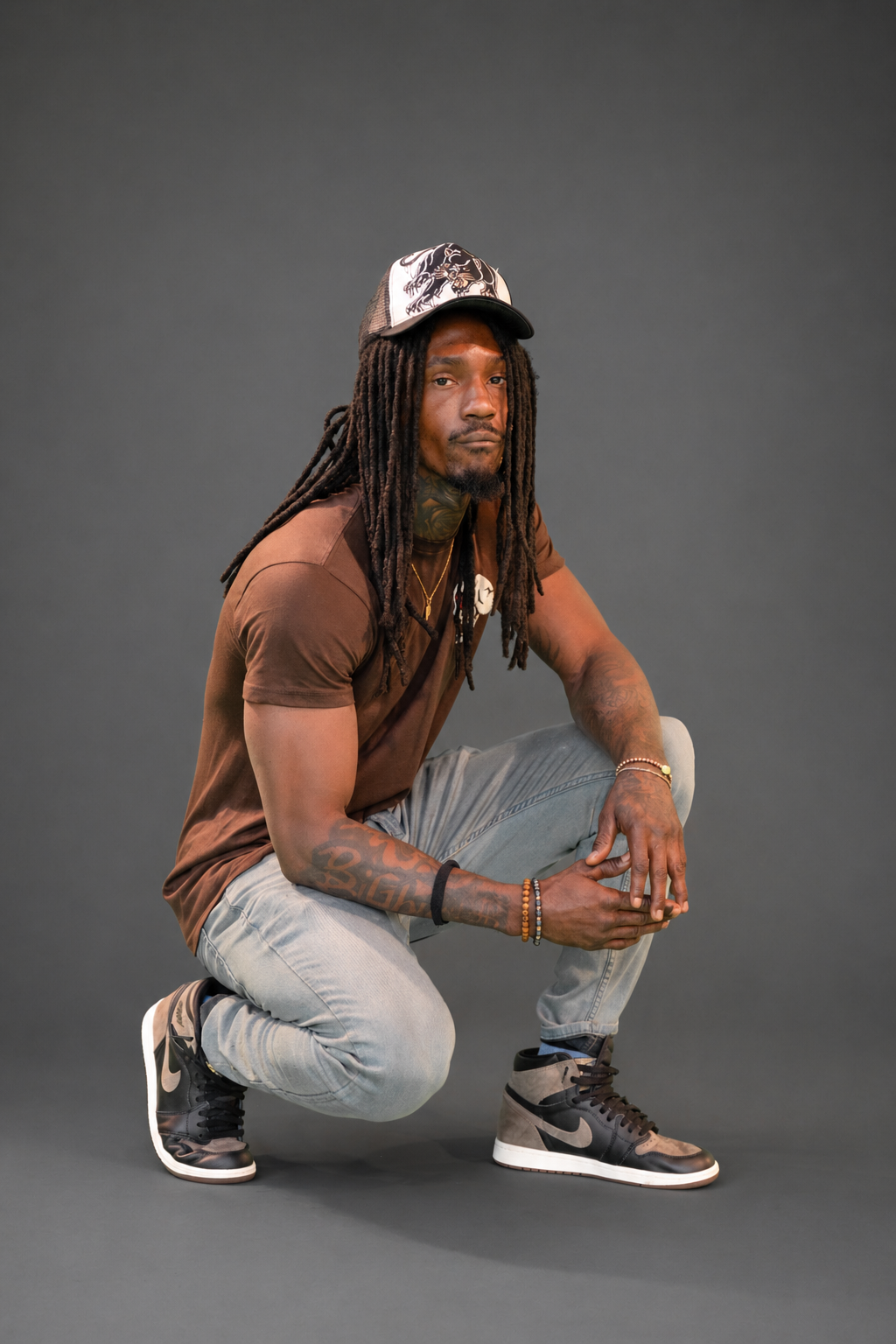 A man with long dreadlocks, tattoos, and a cap crouches on one knee against a grey background.