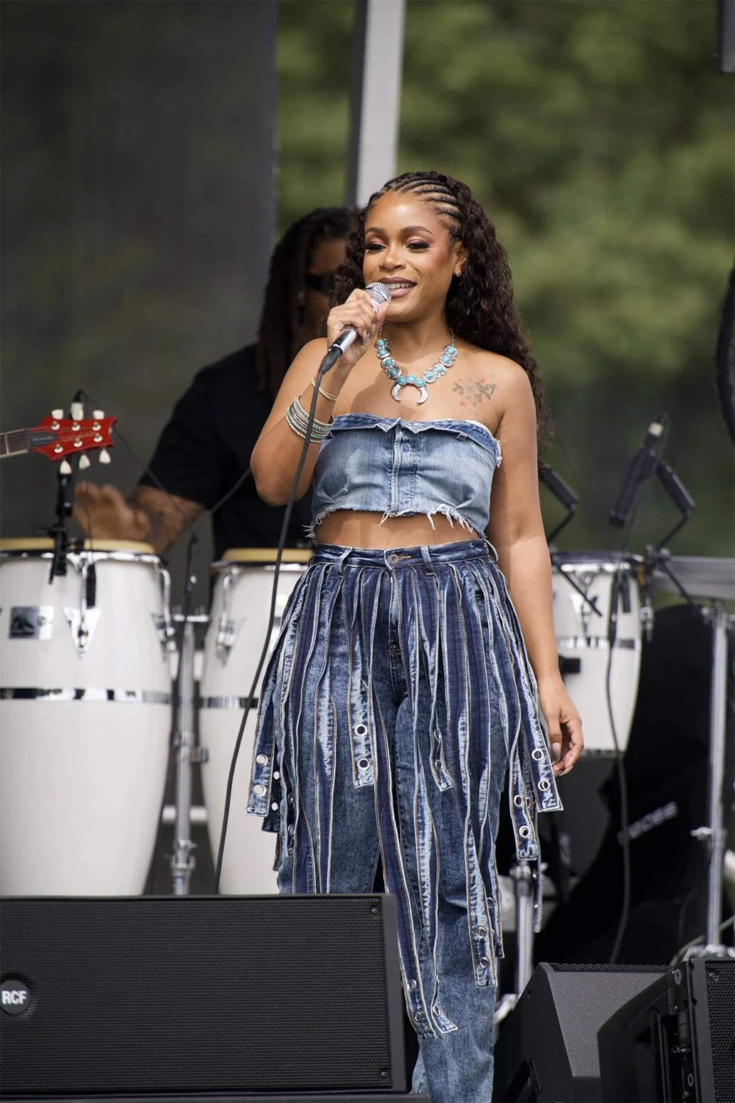 A woman singing into a microphone on stage, wearing a denim crop top and matching striped denim pants, with a musician playing congas behind her.