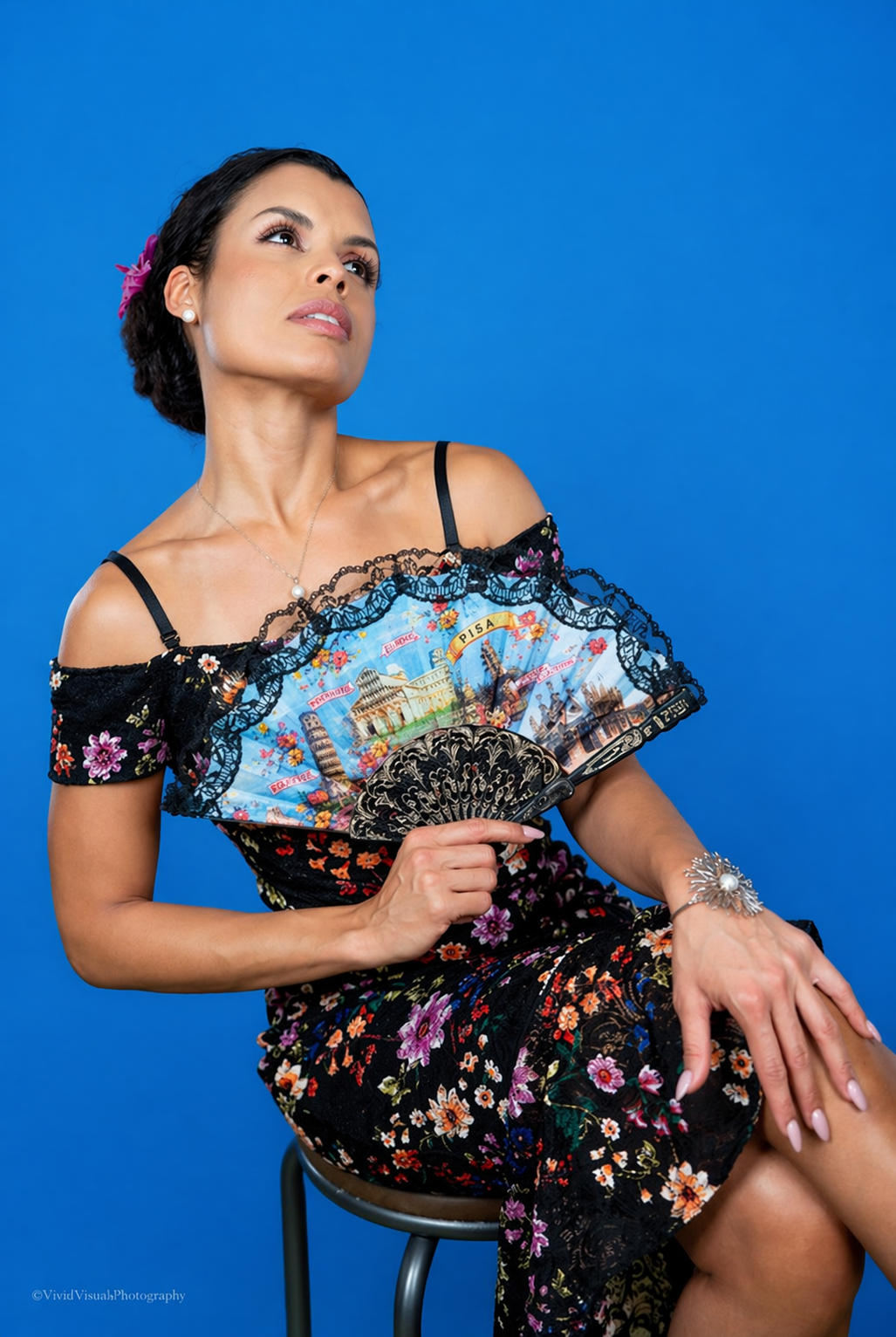 A woman with black hair styled in an updo, wearing a black floral dress, holding a decorated fan, and wearing a jewelry bracelet and pearl earrings, sitting on a stool against a solid blue background.