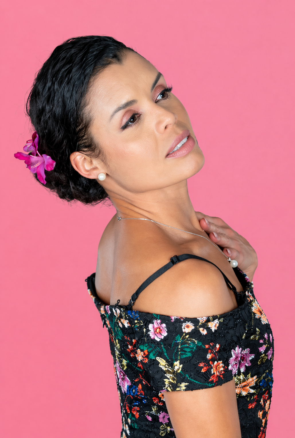 A woman with dark, curly hair adorned with a pink flower, wearing pearl earrings, a pearl necklace, and a floral off-shoulder dress against a pink background.