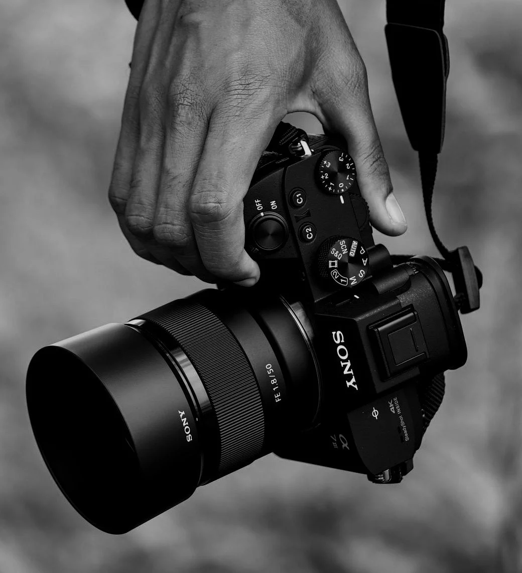 Close-up of a person's hand holding a Sony mirrorless camera with a large lens. The image is in black and white.