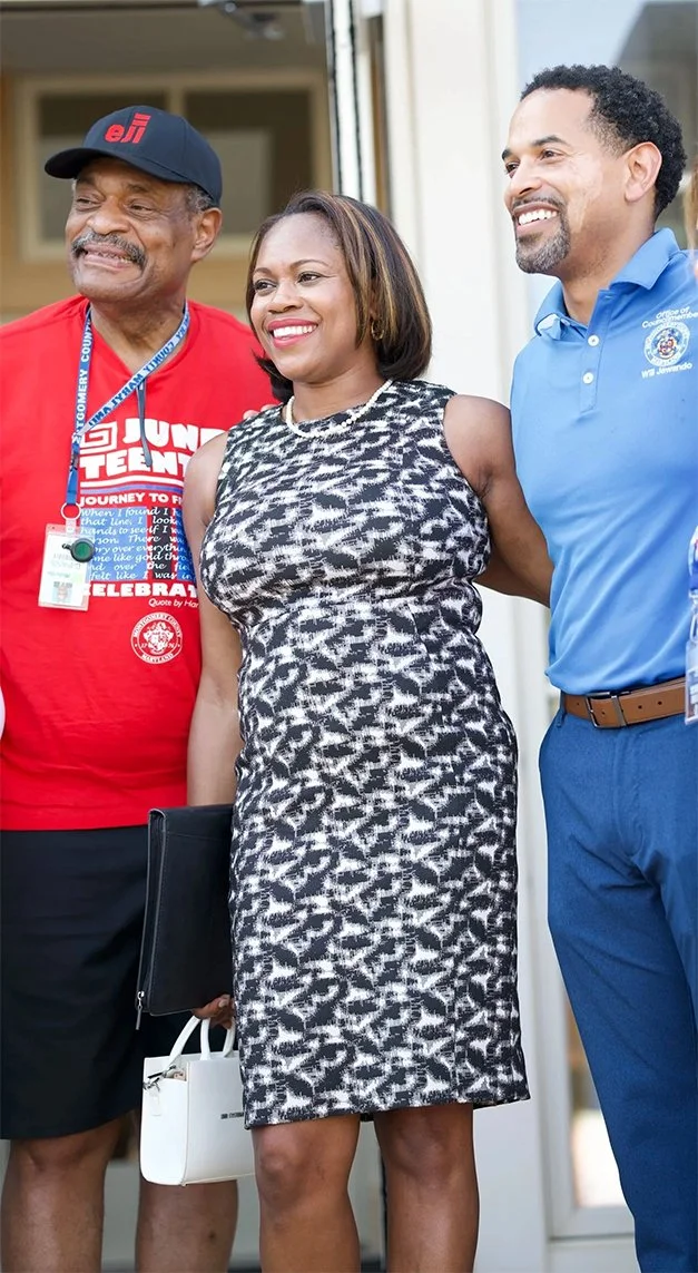 Three people standing together, smiling, outdoors on a sunny day. The woman in the center is wearing a sleeveless black and white patterned dress, holding a white handbag and black folder. The man on the right is in a blue collared shirt and dark pants. The man on the left is wearing a red shirt, a black cap, and a lanyard.