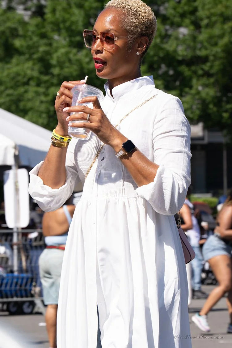 An older woman with short blonde curly hair, wearing sunglasses, a white dress, and gold jewelry, is holding a beverage cup with a straw while outdoors.