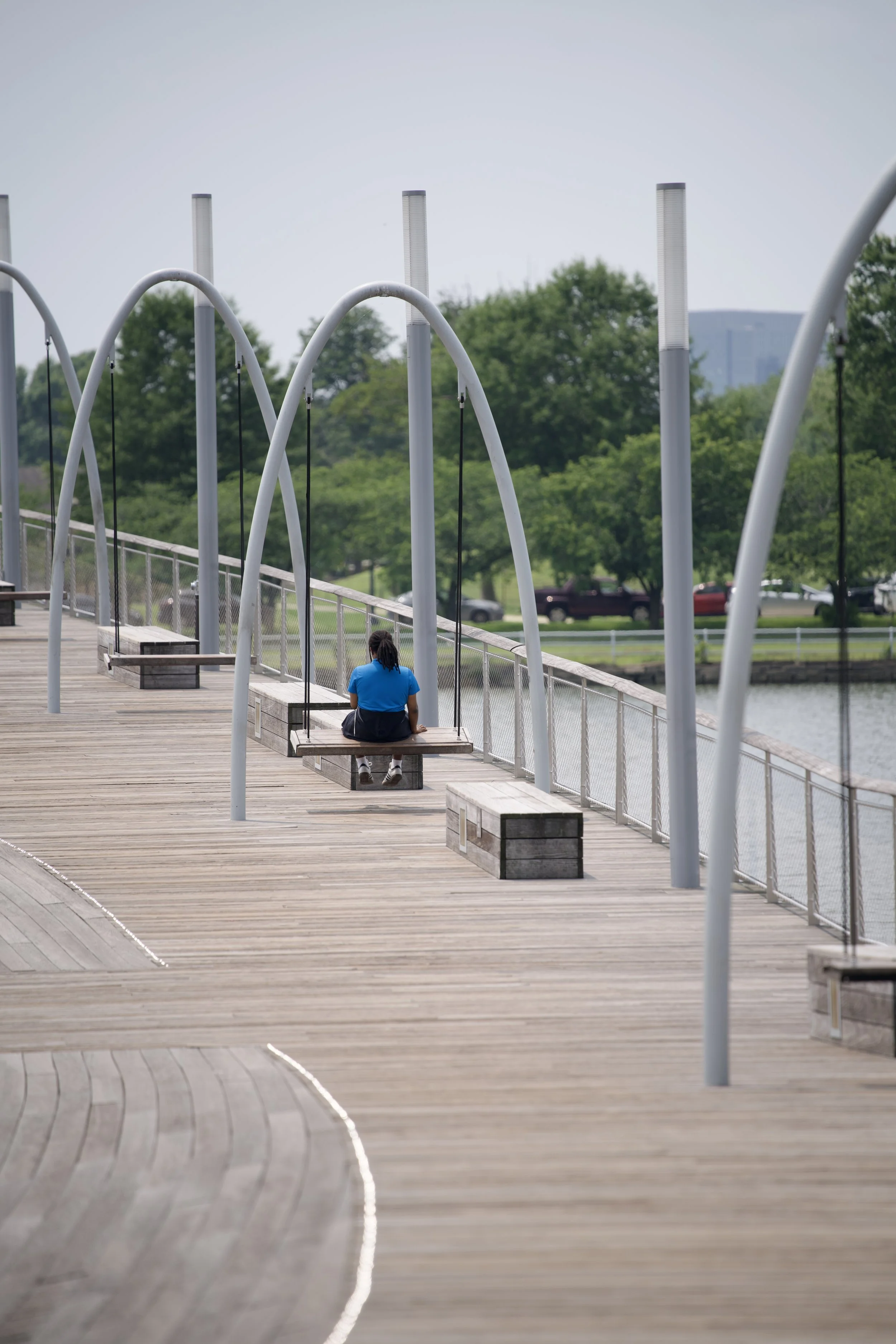 A person sitting alone on a wooden swing bench along a waterfront walkway with arching metal supports, green trees, and parked cars in the background.