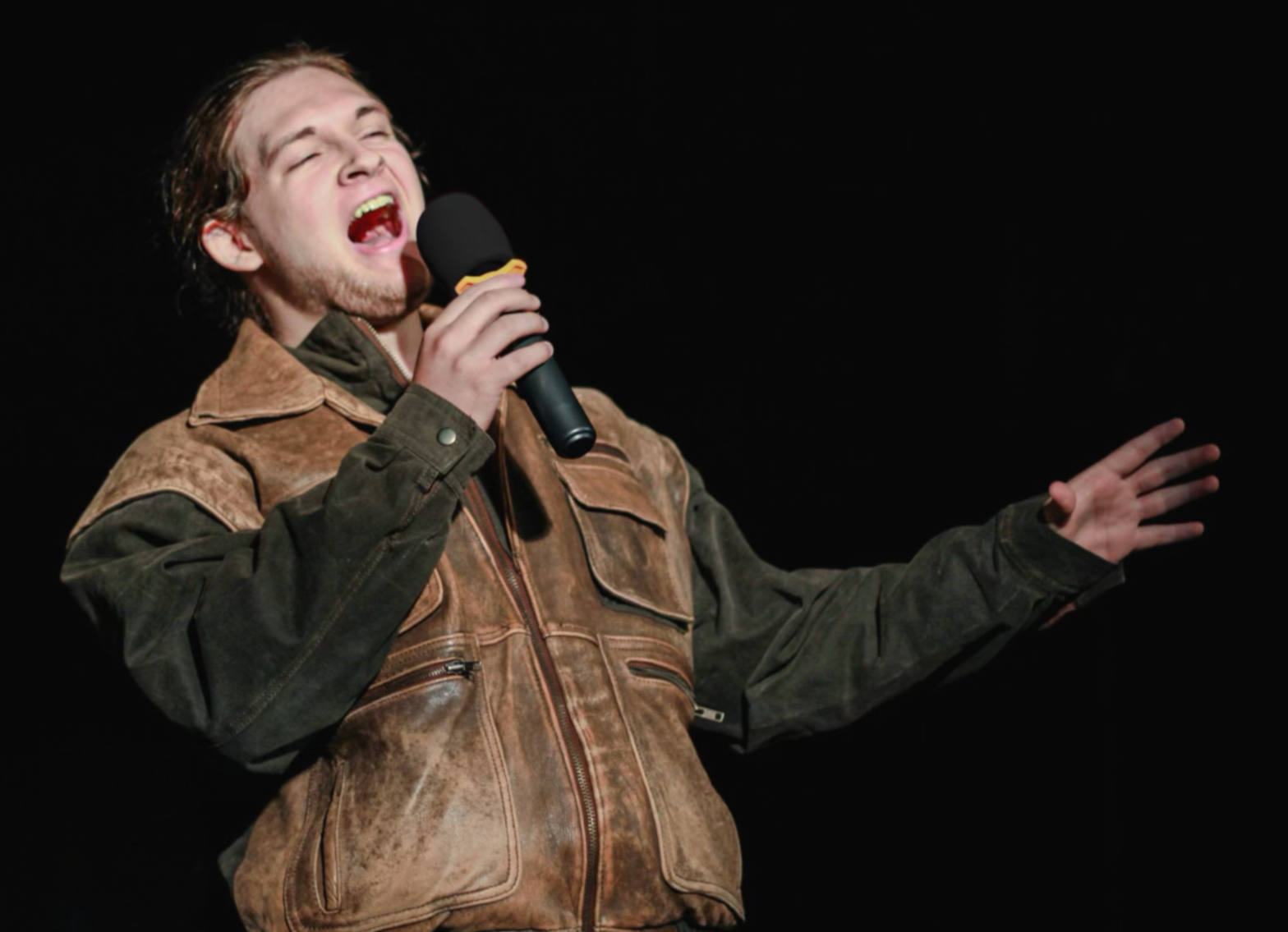 A male performer singing into a microphone on a dark stage, wearing a brown and green jacket with patches.