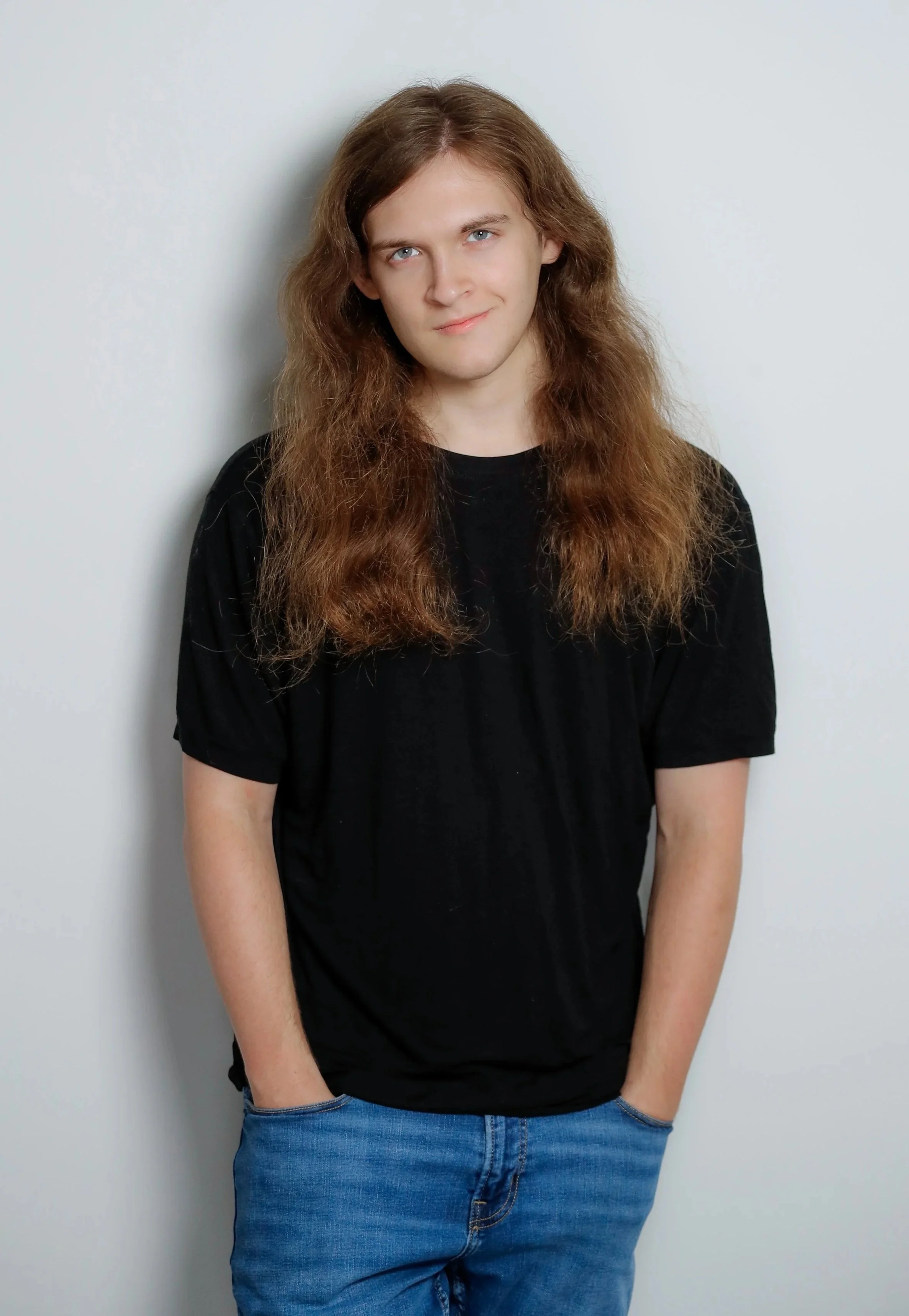 A young man with long wavy brown hair wearing a black T-shirt and blue jeans, standing against a plain white wall, looking at the camera with a slight smile.