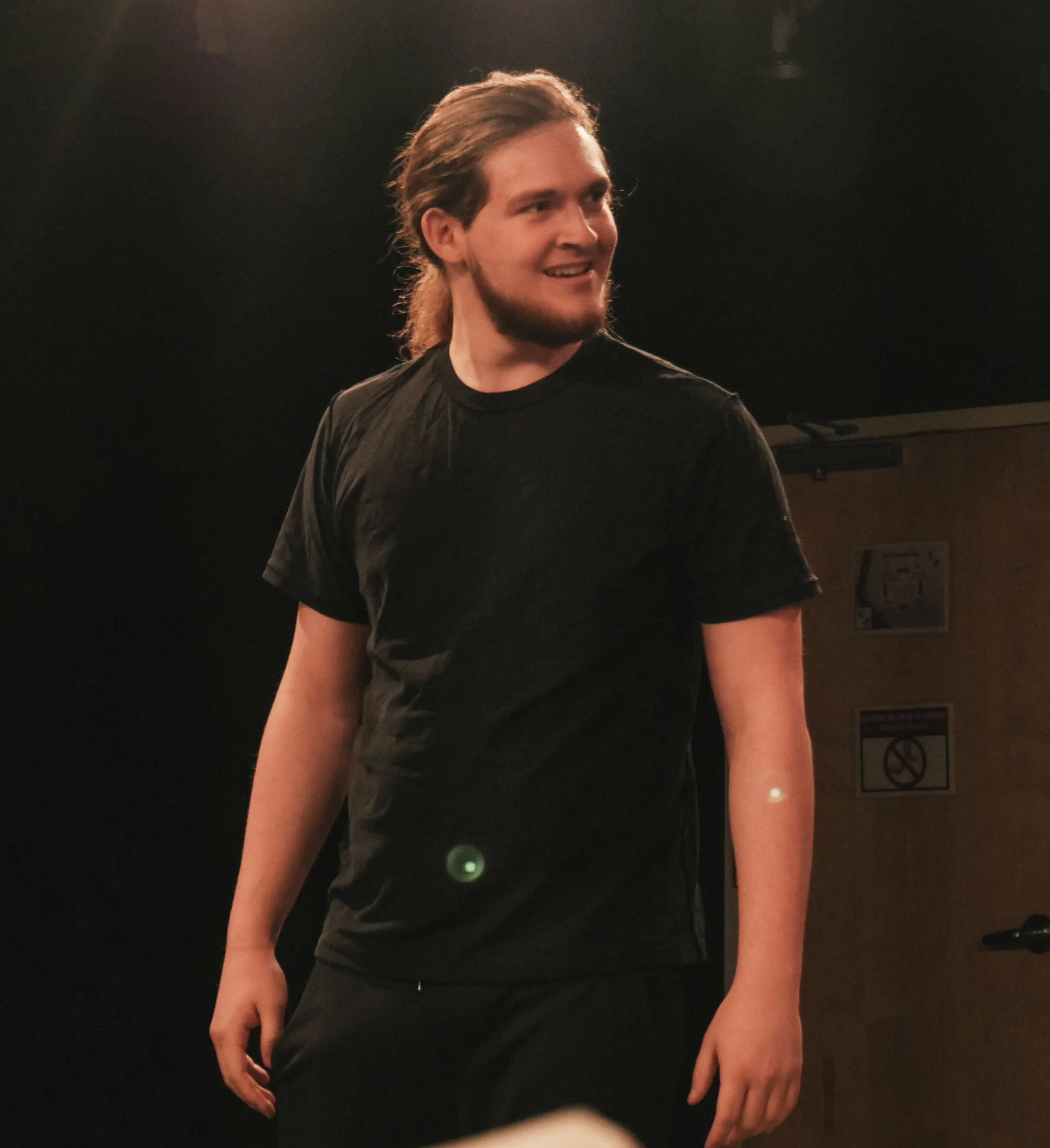 A young man with long hair tied back, wearing a black t-shirt, smiling and standing in a dark room with wooden panels on the wall.