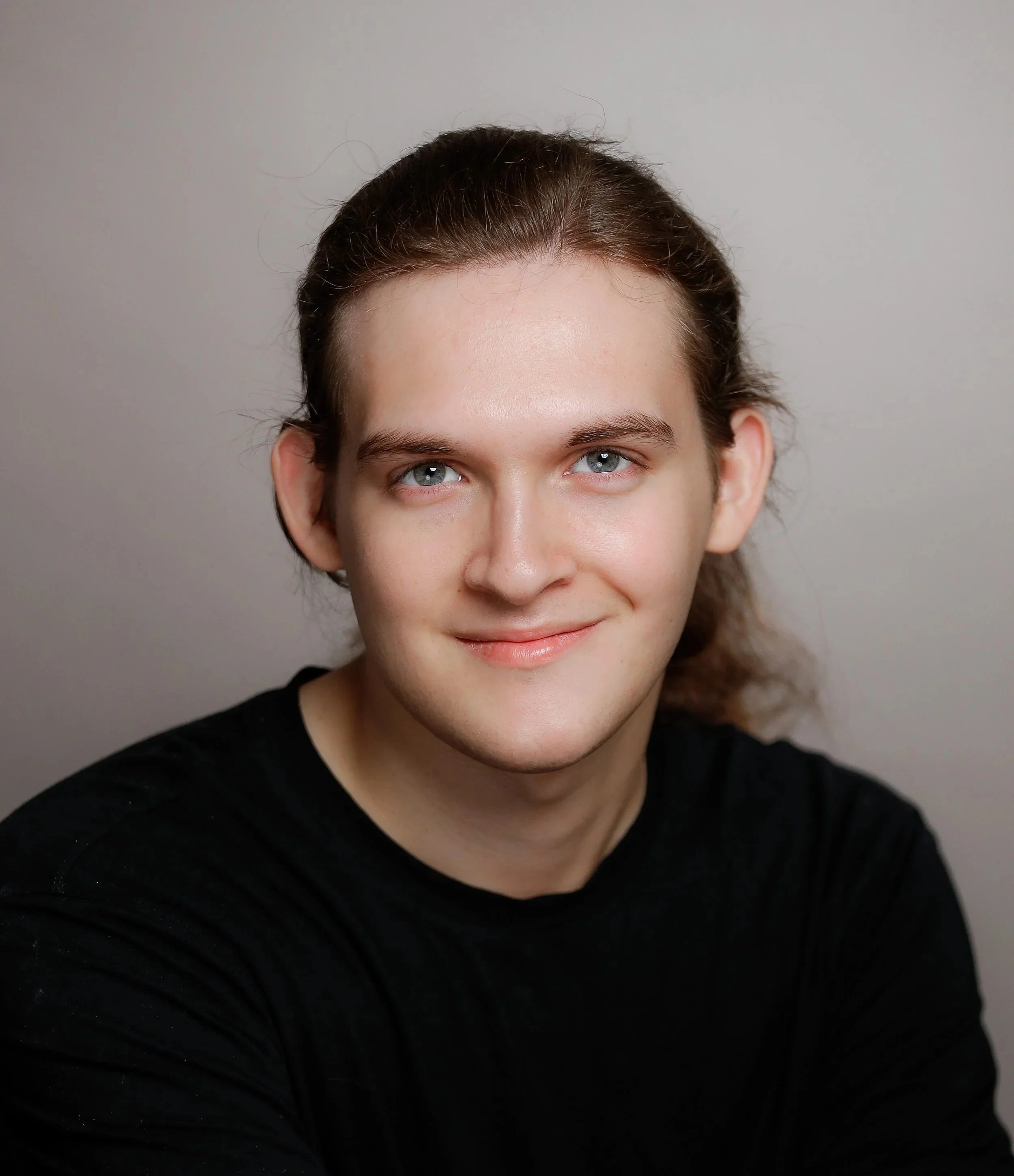 A young man with long brown hair, blue eyes, and light skin, smiling in front of a plain background.