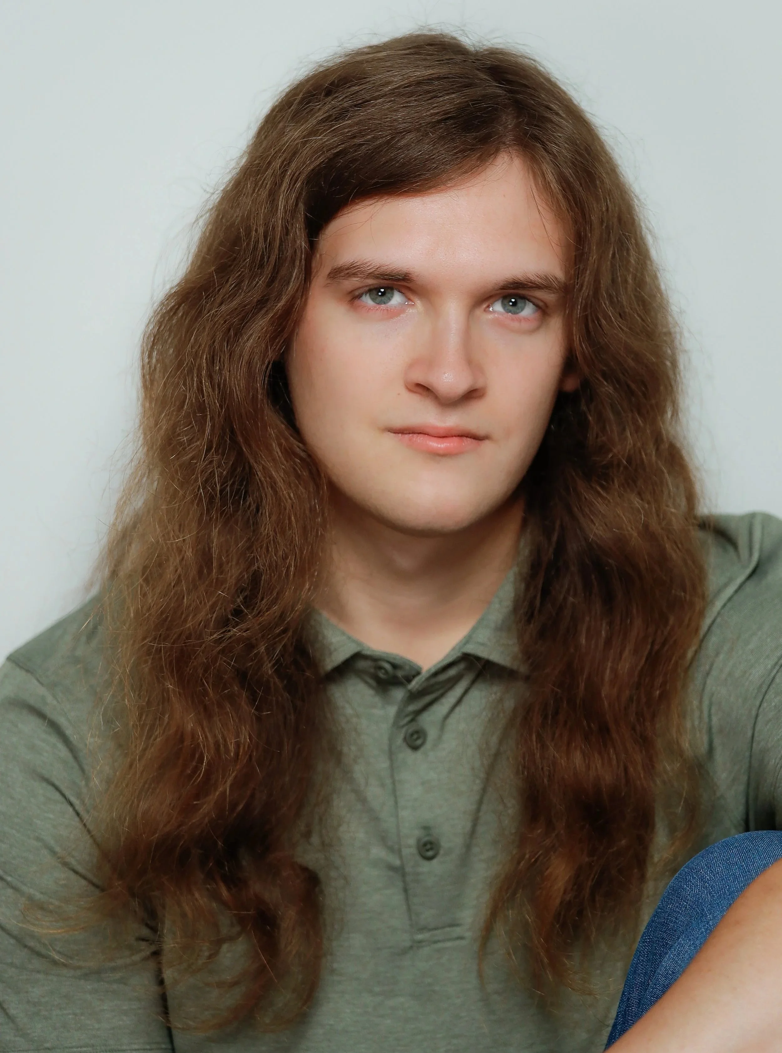 A young man with long, wavy brown hair and blue eyes wearing a green collared shirt, looking at the camera.