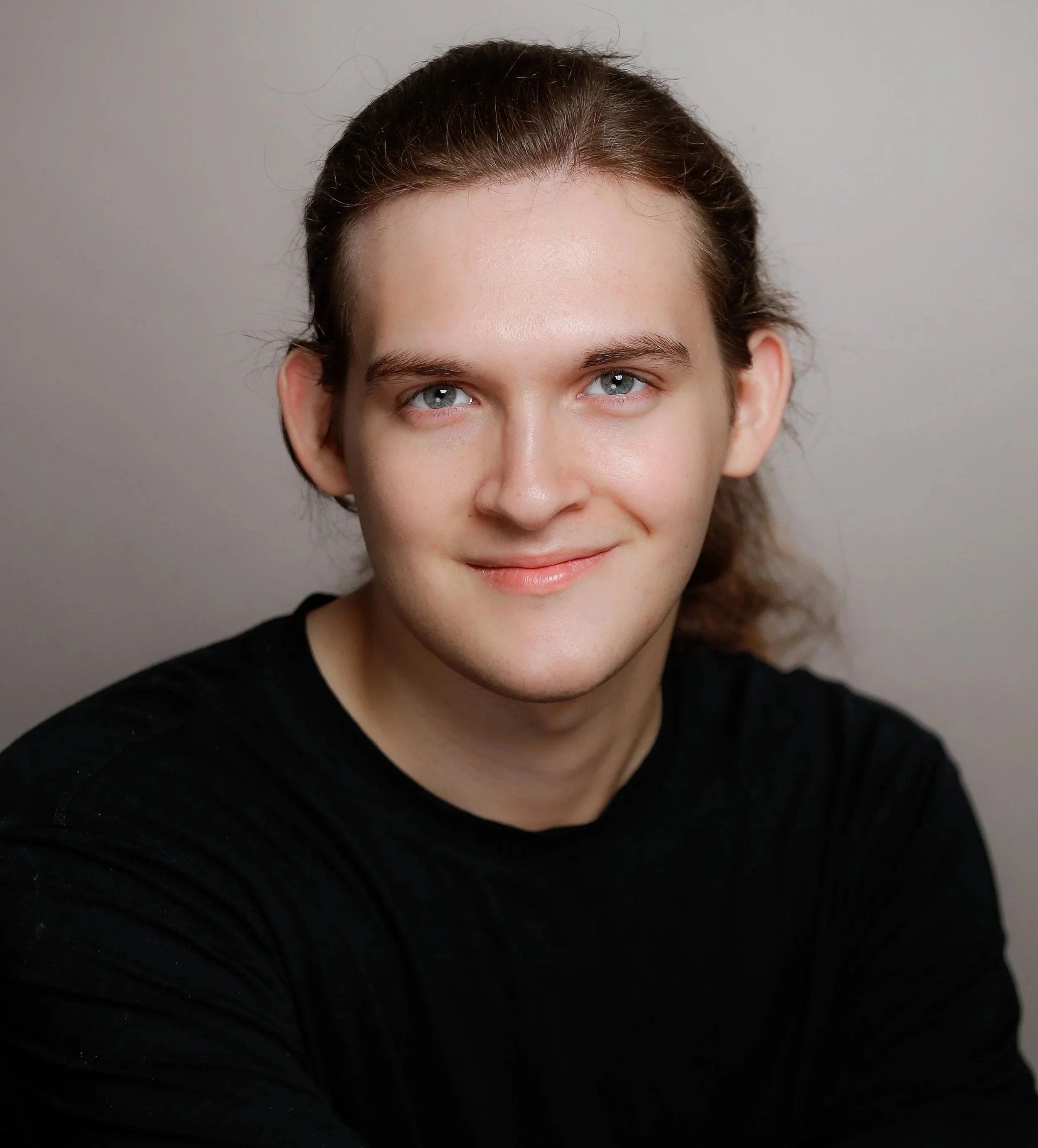 Close-up portrait of a young man with light skin, long brown hair, and blue eyes, smiling and wearing a black shirt, against a neutral background.