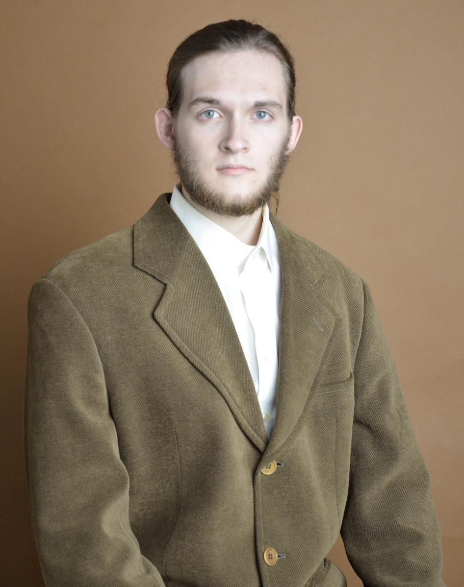 A young man with long hair, a beard, and blue eyes wearing a brown blazer and white shirt, standing against a plain brown background.