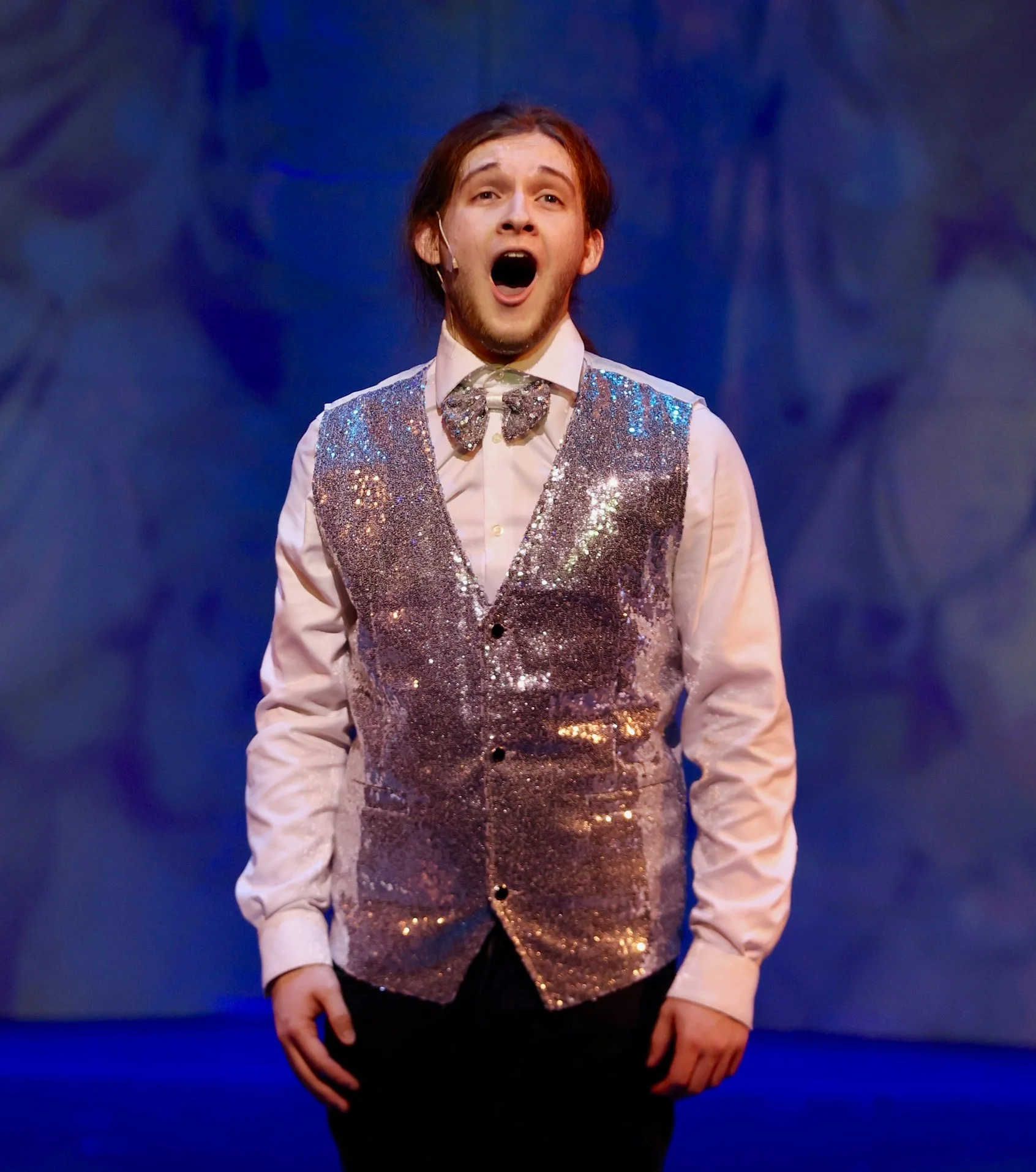 Male performer on stage wearing a sparkly vest and bow tie, singing with a dramatic expression.