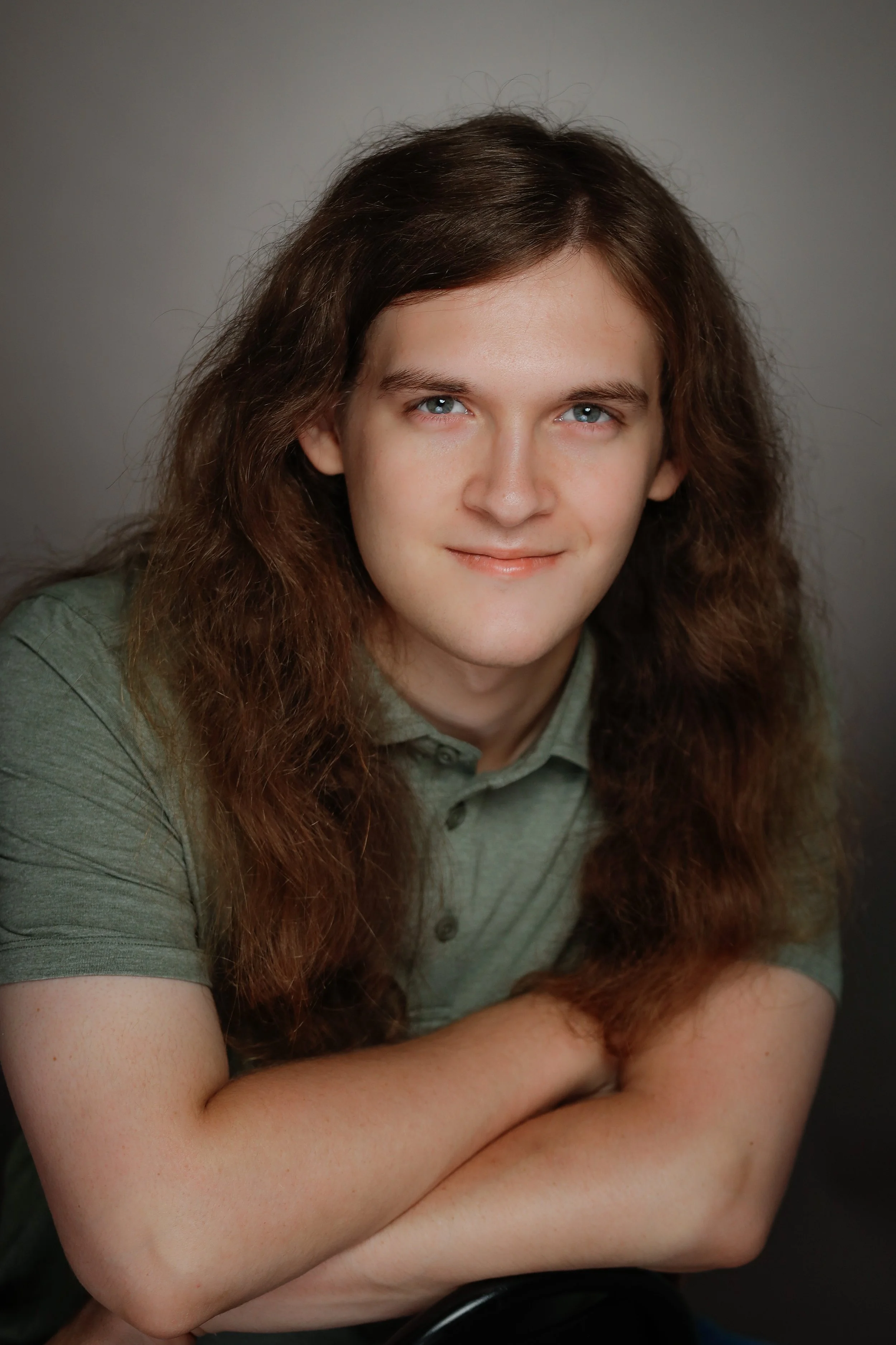 A young man with long, wavy brown hair and blue eyes, wearing a green collared shirt, smiling softly with arms crossed, posed against a neutral background.