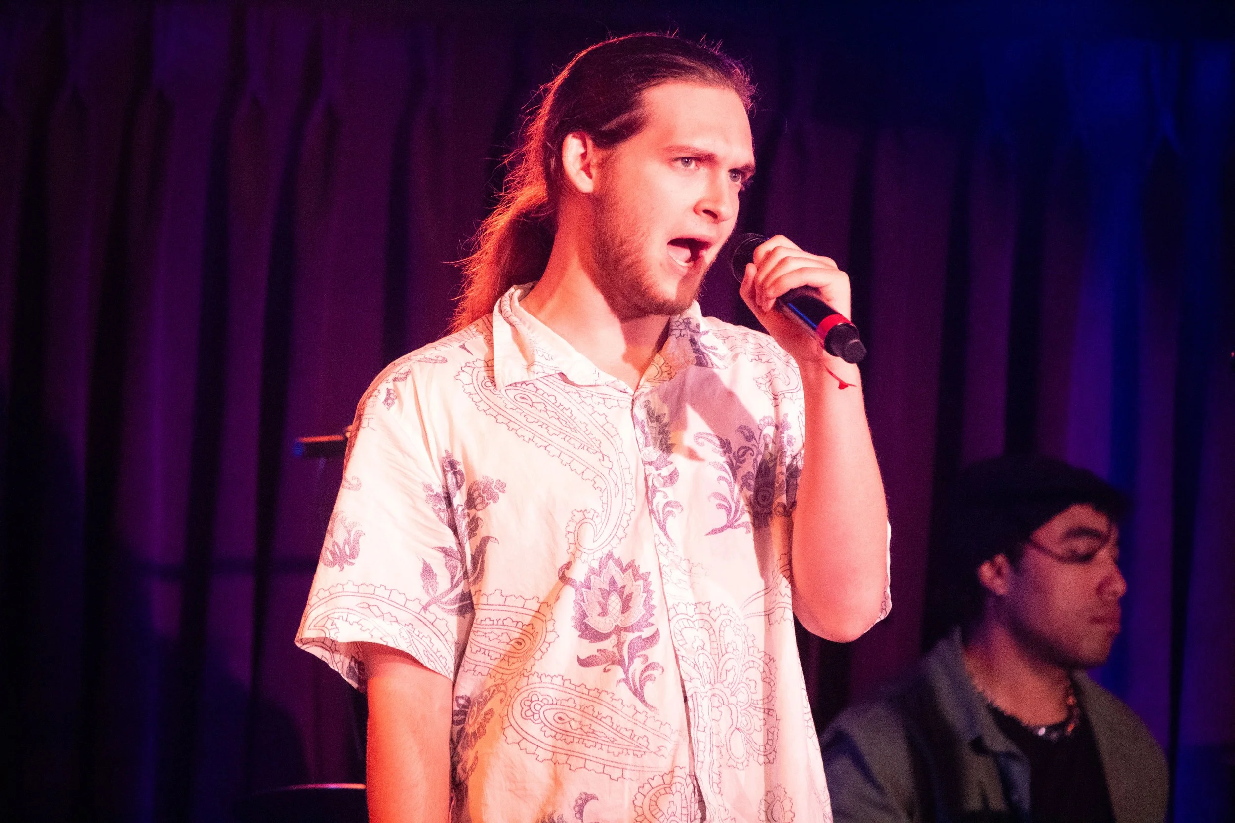 A young man singing into a microphone on stage, wearing a light-colored patterned shirt, with a dark curtain background and another person in the background.